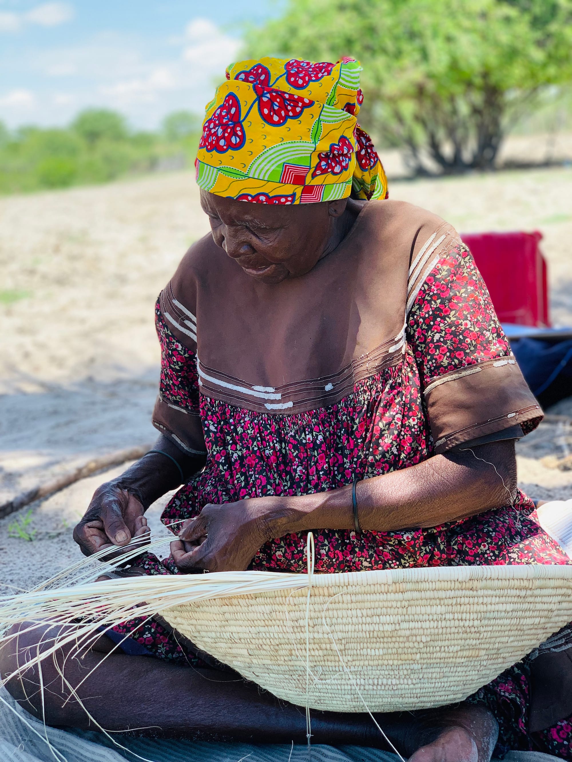 An elderly woman in a bright patterned headscarf and floral dress sits on sandy ground weaving a large, shallow coiled basket from pale strips of palm leaf