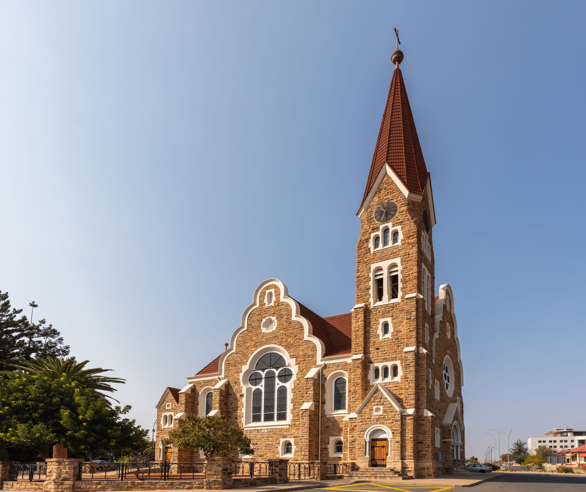 A sandstone church with white-trimmed gables, arched windows and a tall tower topped by a red-tiled spire and a cross, under a clear pale blue sky