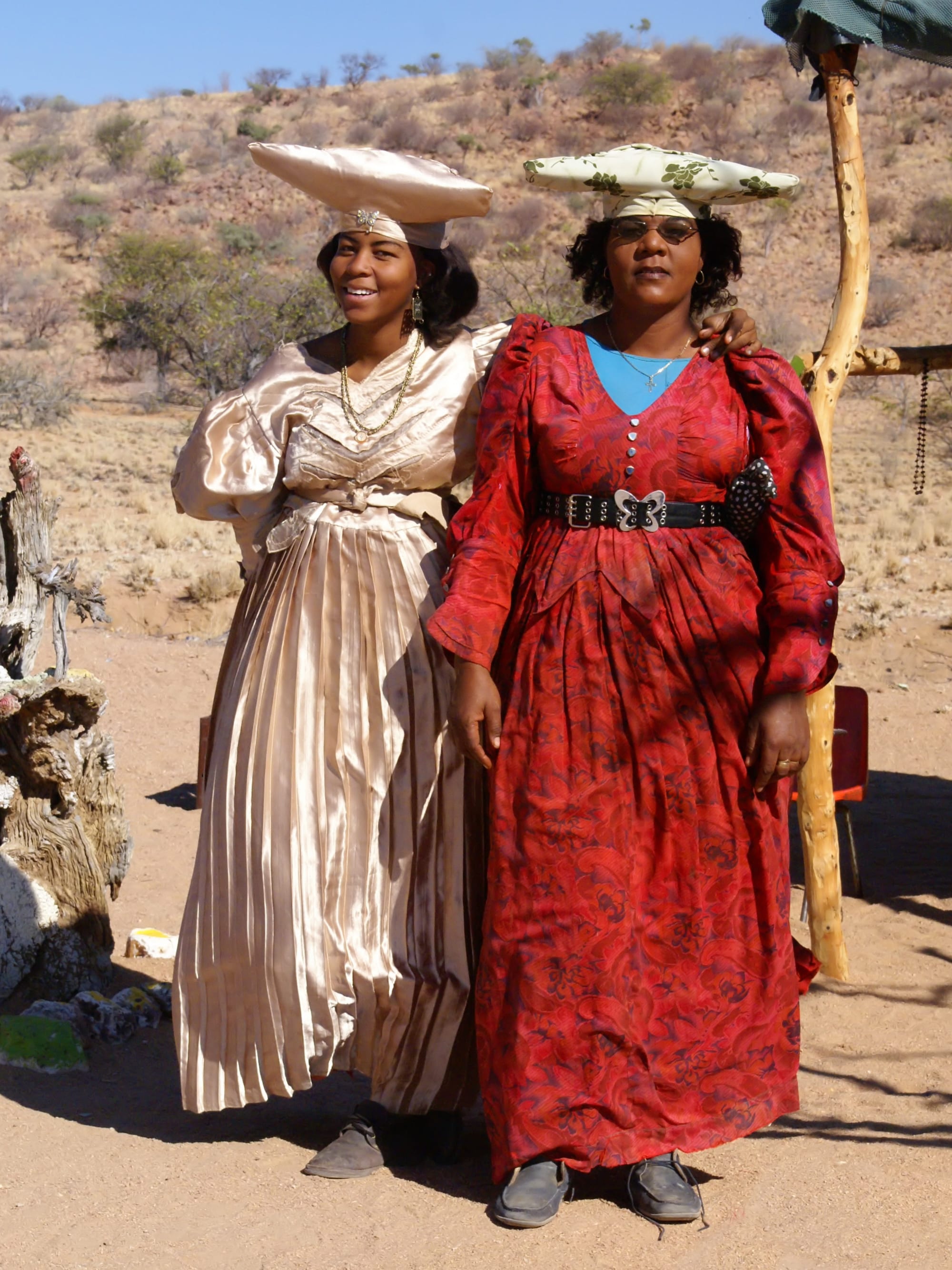 Two women stand side by side on sandy ground in front of dry scrub-covered hills, one in a long pleated champagne satin dress and the other in a long red patterned dress, both wearing wide horizontal headdresses