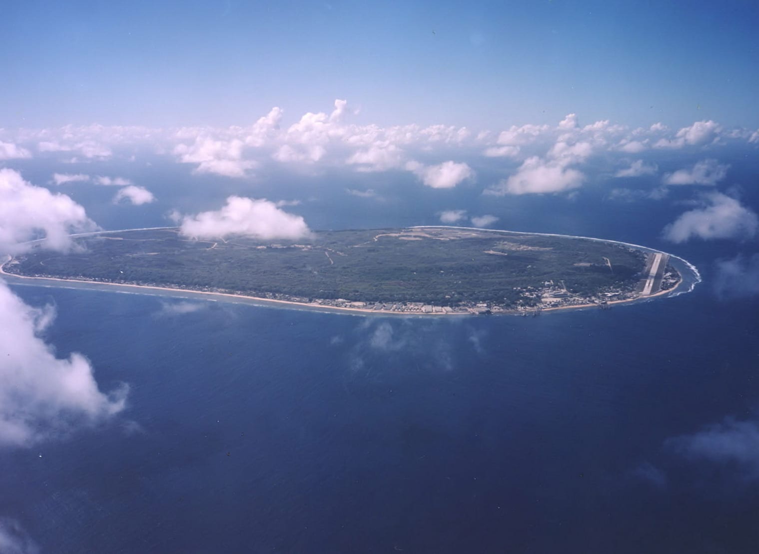 A small coral island with a narrow green coastal ring around a pale, scarred interior plateau
