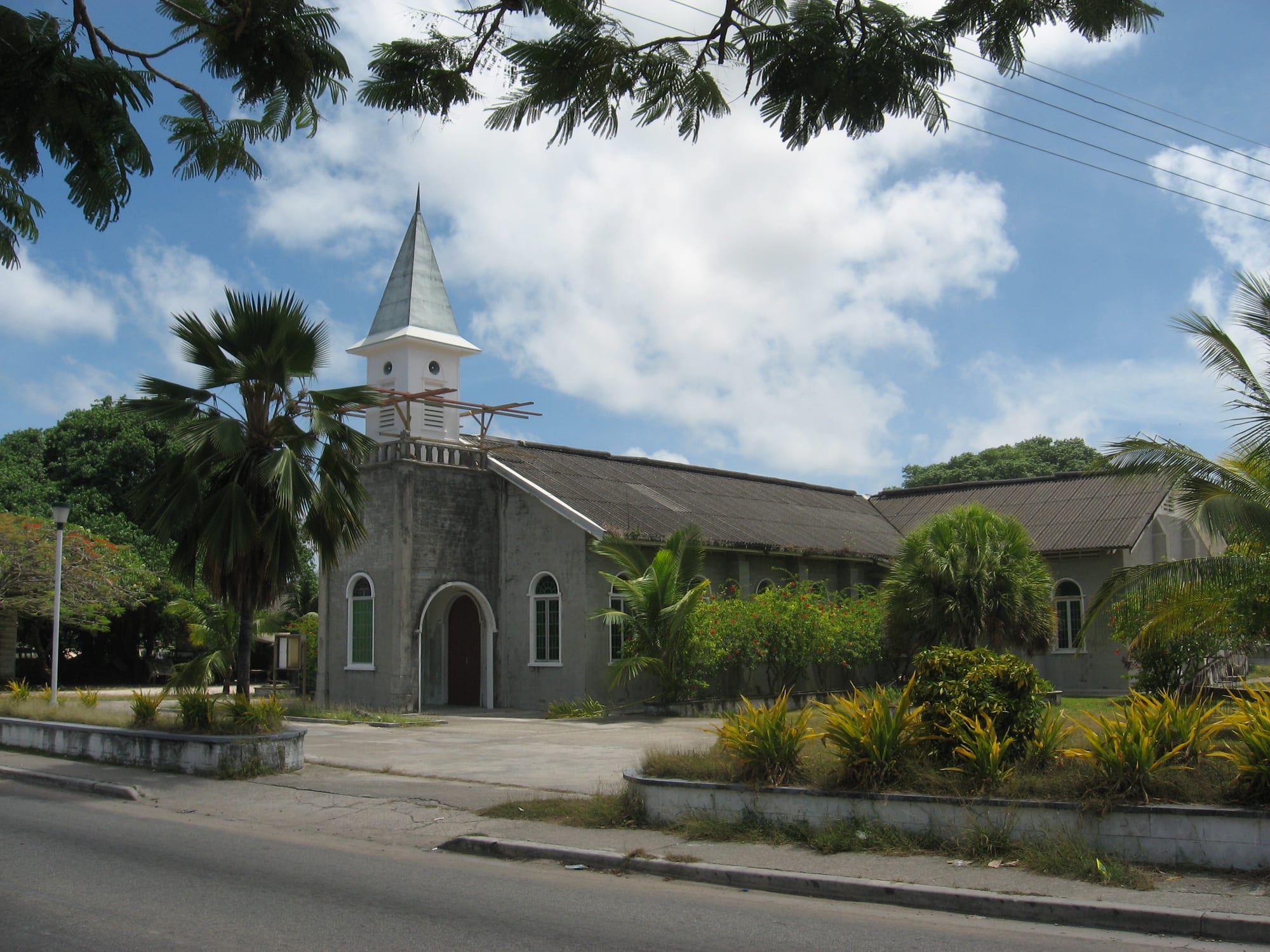 A grey concrete church with arched windows, a pointed white bell tower and a long low roof, set behind flower beds and palms beside a road under a blue sky with white clouds