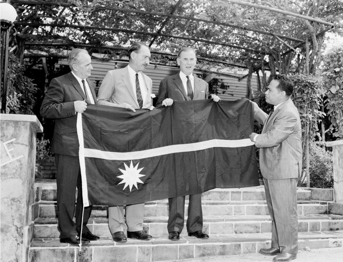 A black and white photograph of three men in suits holding up a large flag with a horizontal stripe and a twelve-pointed star, while a fourth man in a suit on the right holds the other end, on stone steps beneath a vine-covered pergola