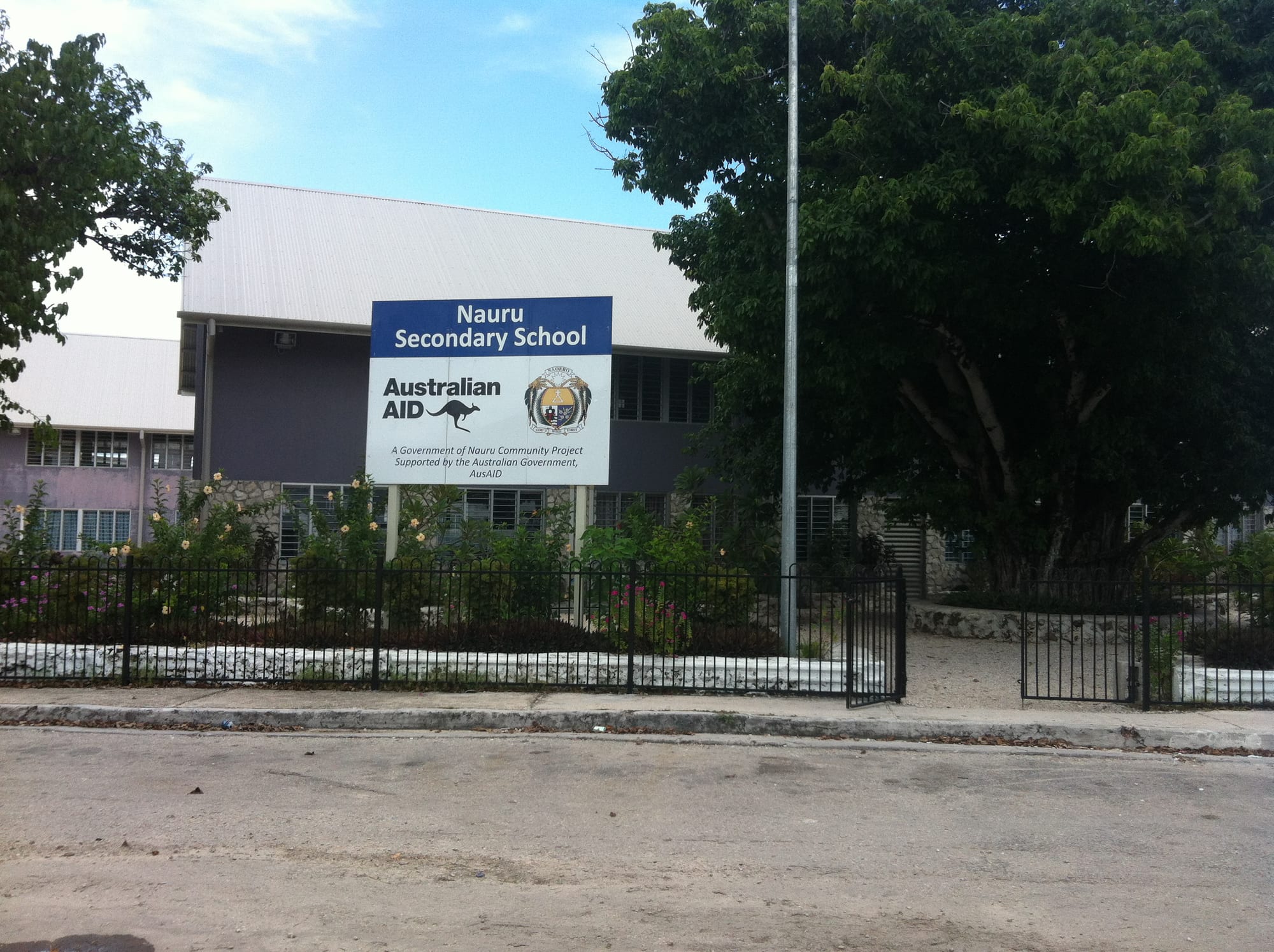A two-storey school building with a sign reading Nauru Secondary School, behind a fence and garden