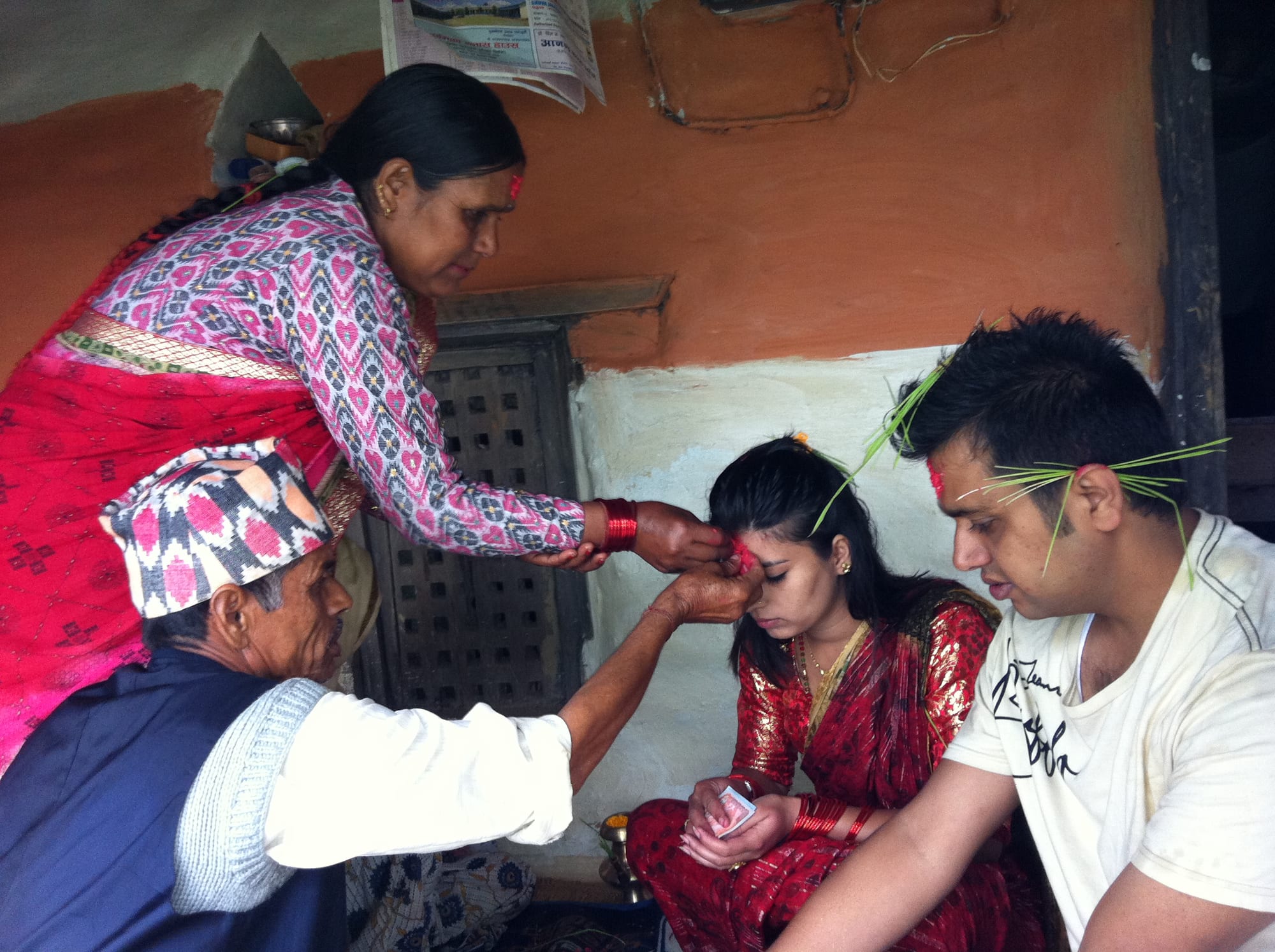 An older woman and an older man in a patterned cap press red tika onto the forehead of a young woman in a red sari, while a young man beside her sits with green jamara shoots tucked into his hair, in a room with an orange-painted wall