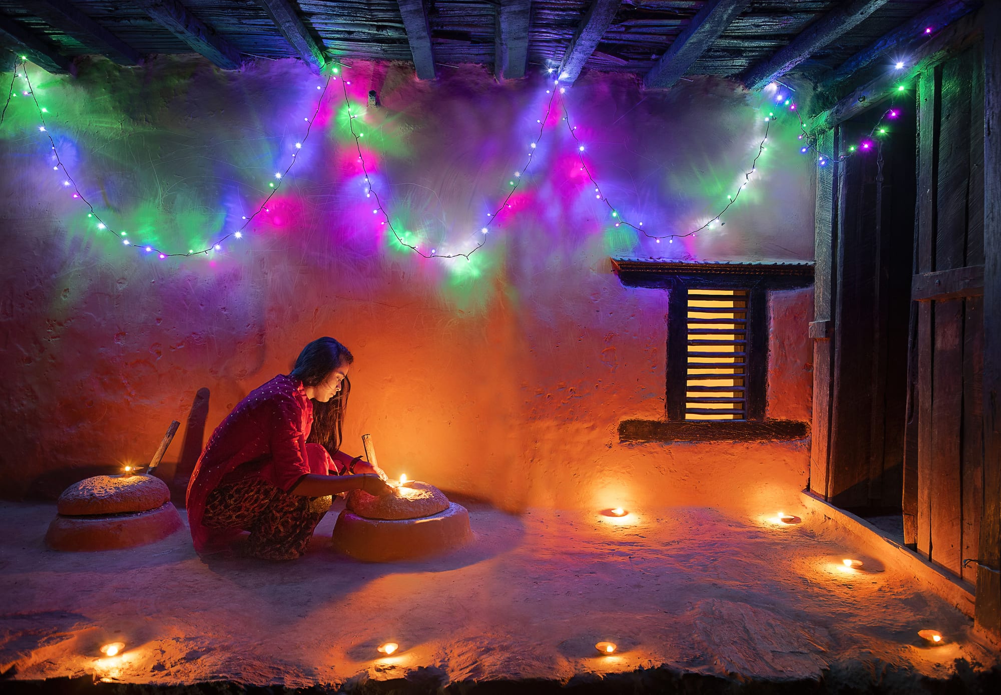 A young woman in a red shawl crouches to light small oil lamps in the mud-plastered porch of a traditional house, with more lamps glowing on the floor and strings of coloured lights hanging from the wooden ceiling