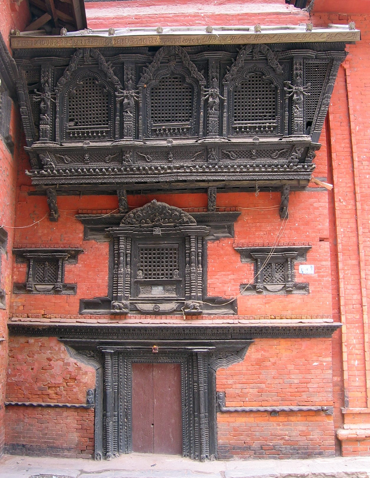 A red-brick house front with a wide projecting carved wooden window with three latticed openings above a smaller carved window and an ornately framed wooden doorway