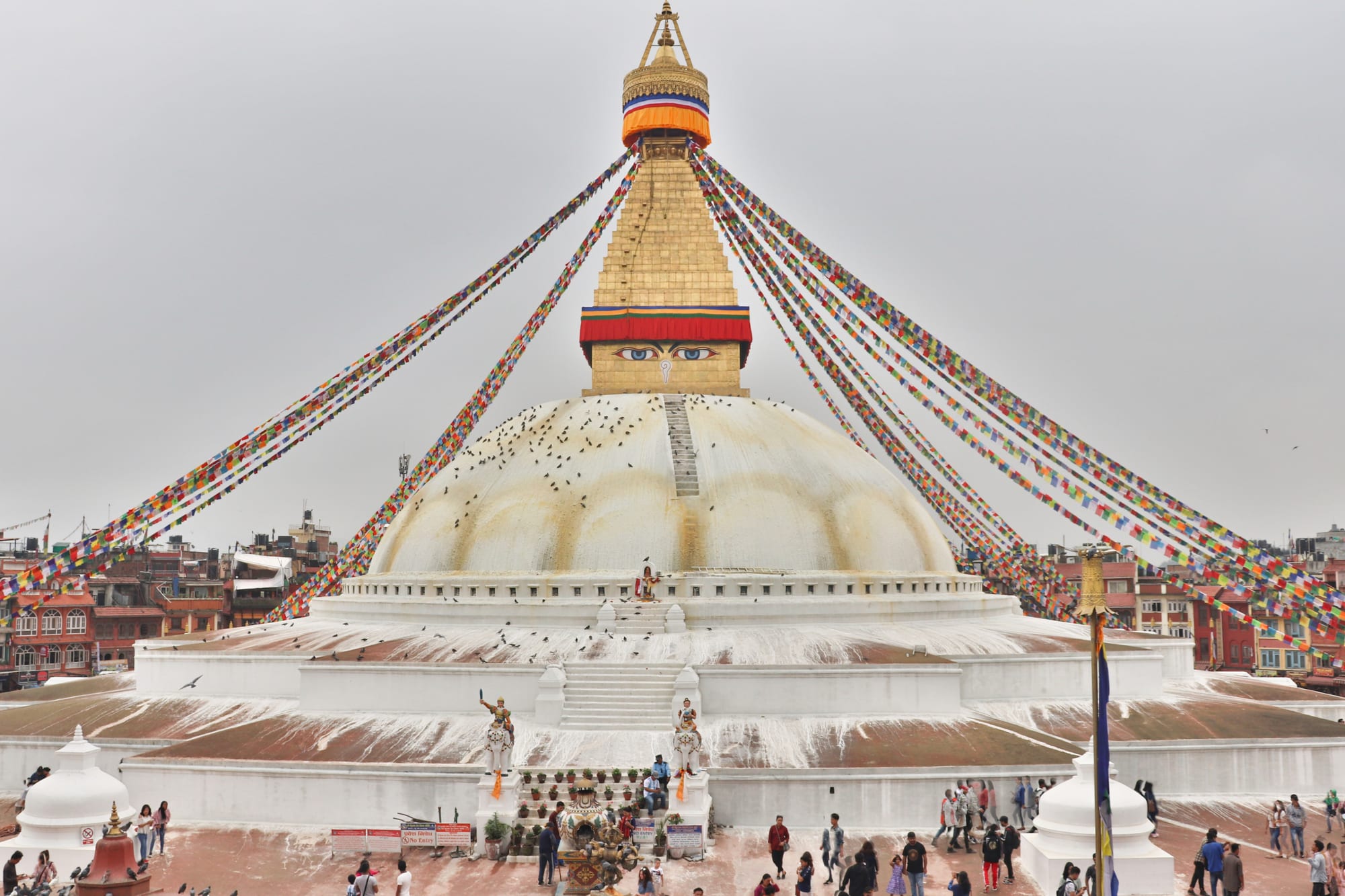 A great white domed stupa topped by a gilded square tower painted with a pair of eyes, with long strings of coloured prayer flags running down from its spire and visitors walking around its base