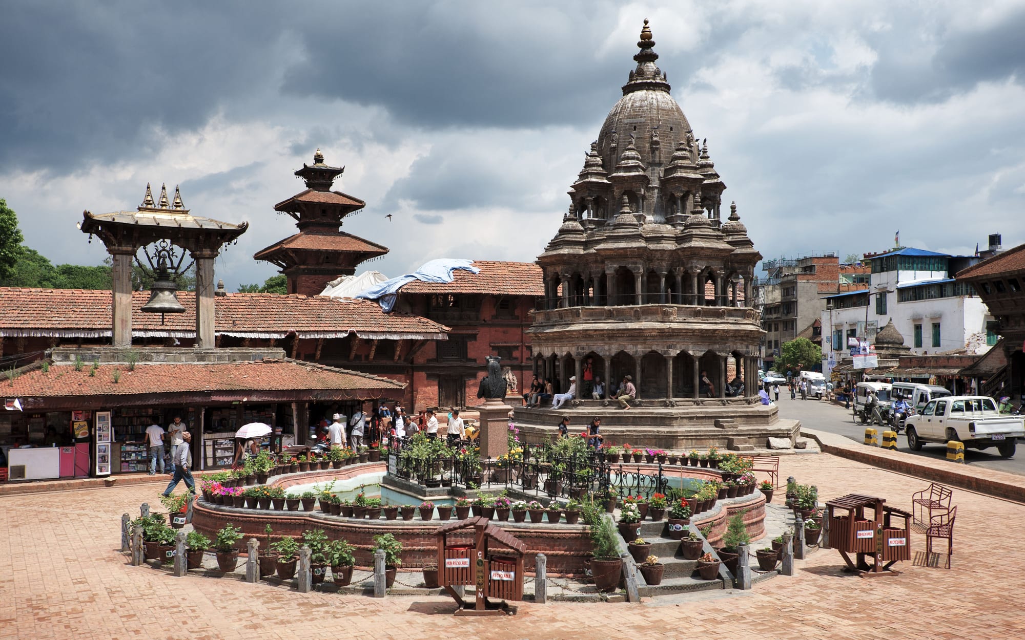 A domed stone temple, a large bell under a pavilion and red-brick palace buildings around a paved square in Patan