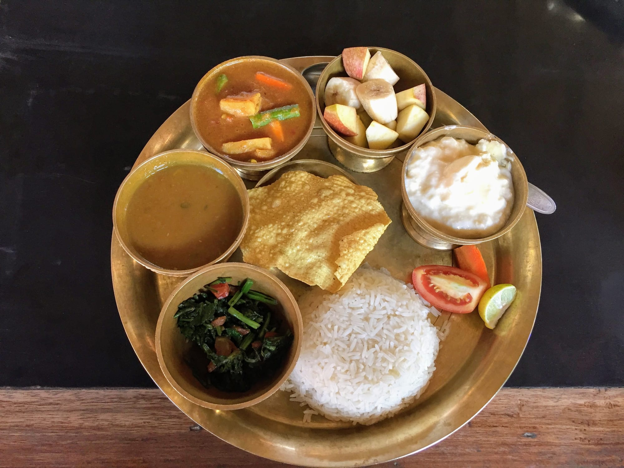 An overhead view of a round brass thali holding rice, lentil soup, vegetable curry, greens, a papad and fruit