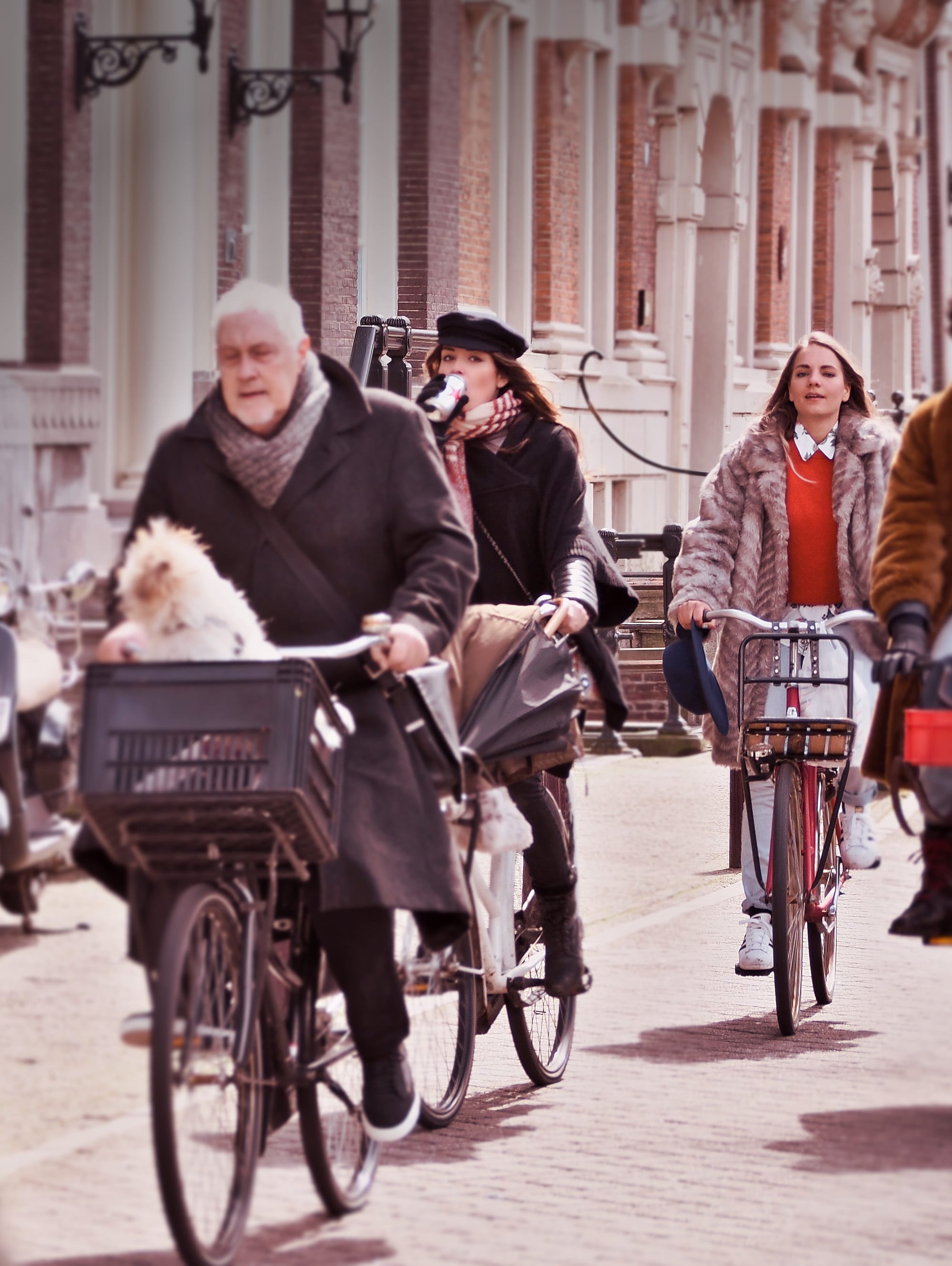 Several cyclists in coats and scarves riding bicycles along an Amsterdam street lined with ornate brick buildings