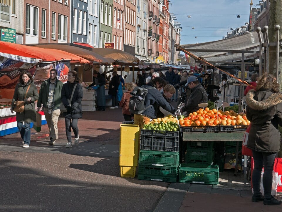 Shoppers walking past a fruit stall stacked with crates of oranges and limes at a sunny outdoor street market
