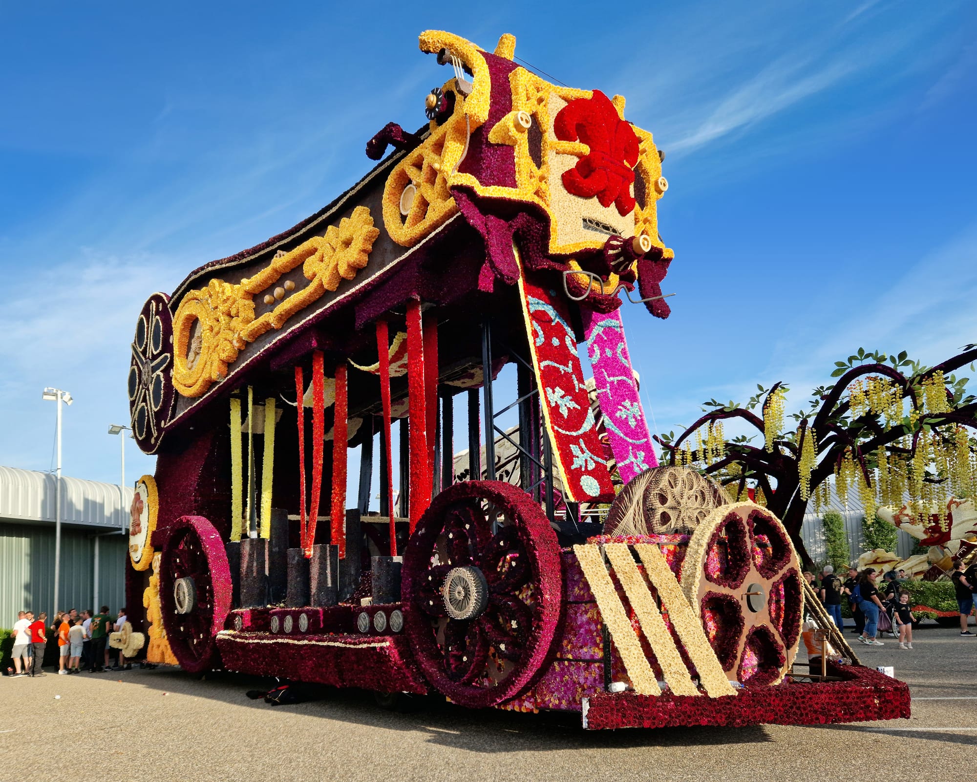 A towering parade float shaped like an ornate carriage, covered entirely in red, purple, yellow and cream flowers, standing on a tarmac square under a blue sky with spectators nearby