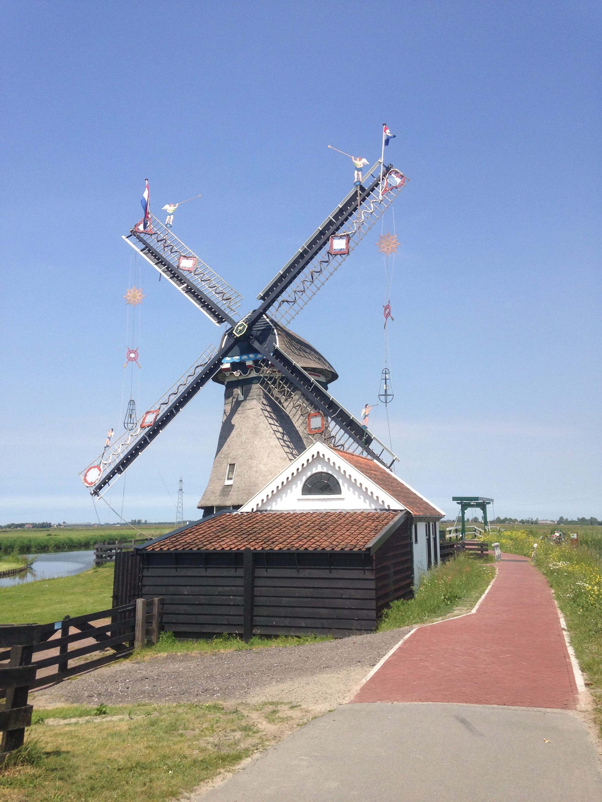 A thatched windmill with dark sails hung with small flags, figures and decorations, standing beside a brick cycle path, a wooden shed and a canal under a clear blue sky