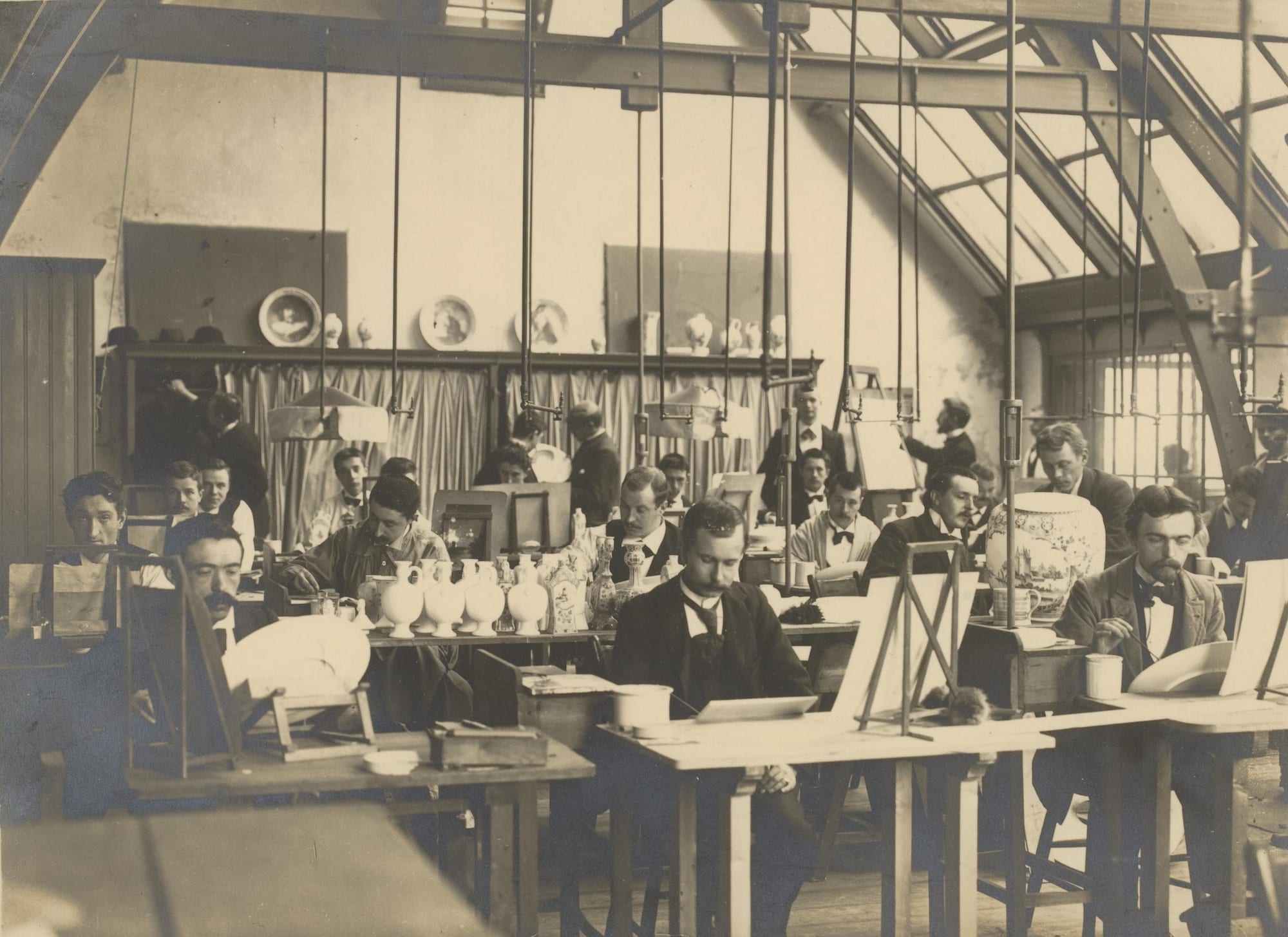 An old sepia photograph of a large studio under a glass roof, where rows of men in suits sit at wooden desks painting vases, jugs and plates