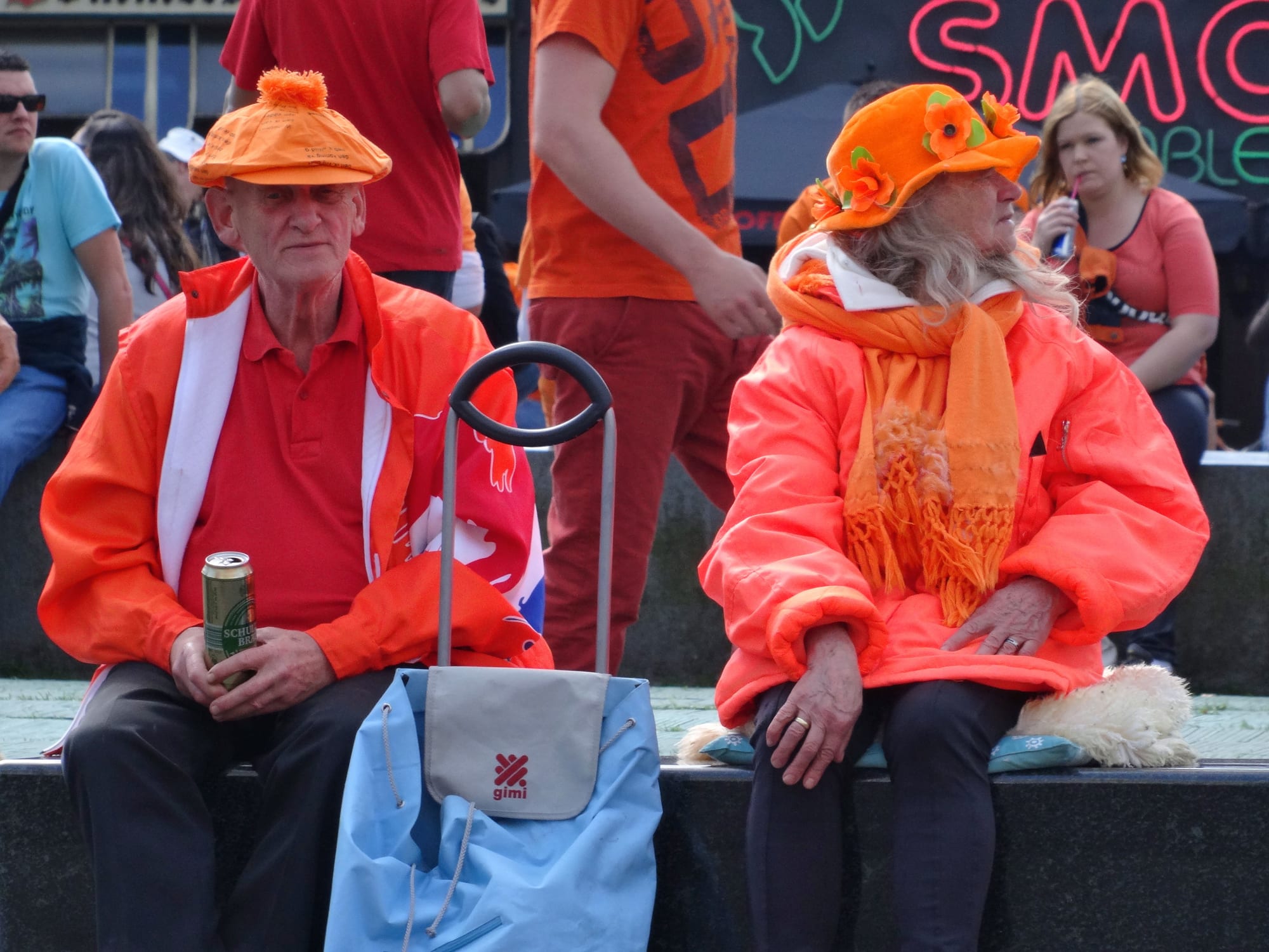 An older man and woman dressed head to toe in bright orange jackets, hats and scarves sit on a stone ledge, with other people in orange behind them