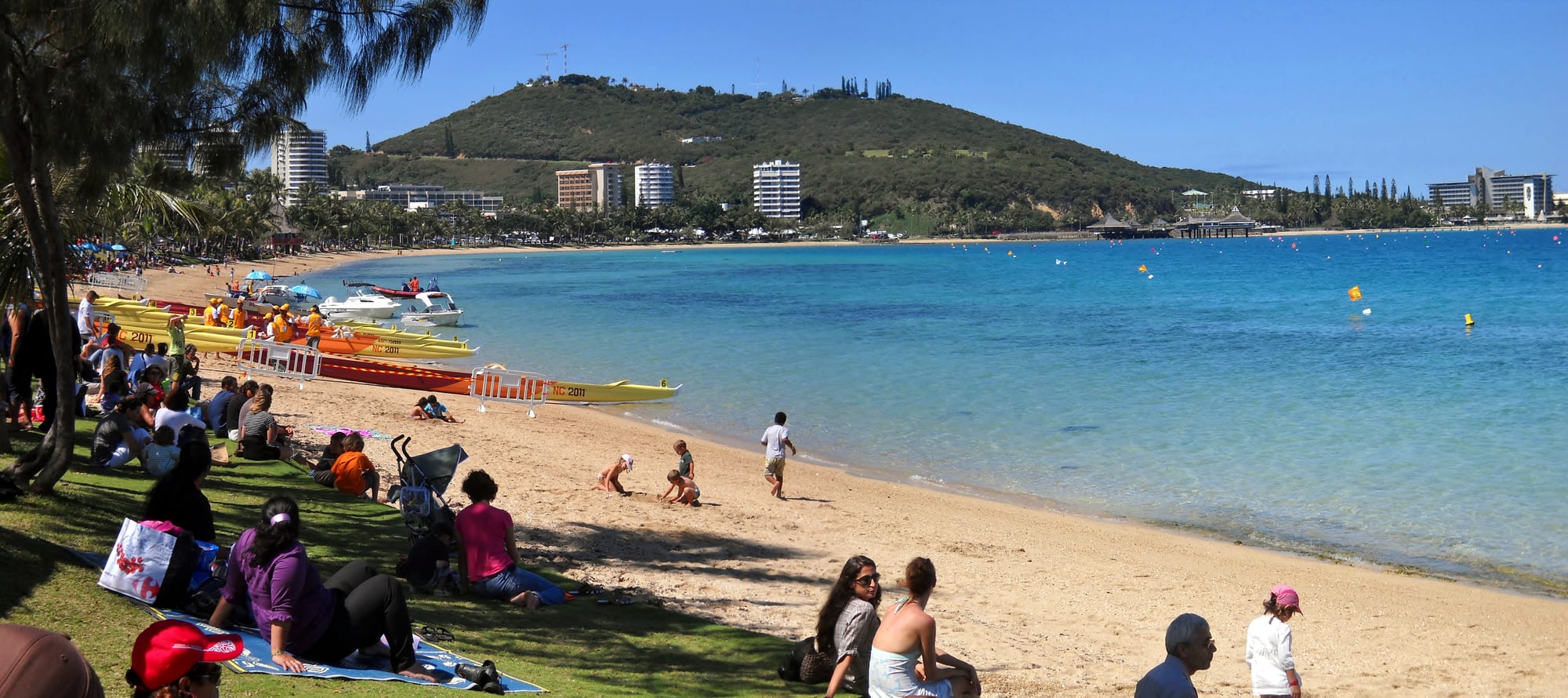 Families relaxing on the sand at Anse Vata beach in Noumea, with outrigger canoes pulled up on the shore and a hill and city skyline behind