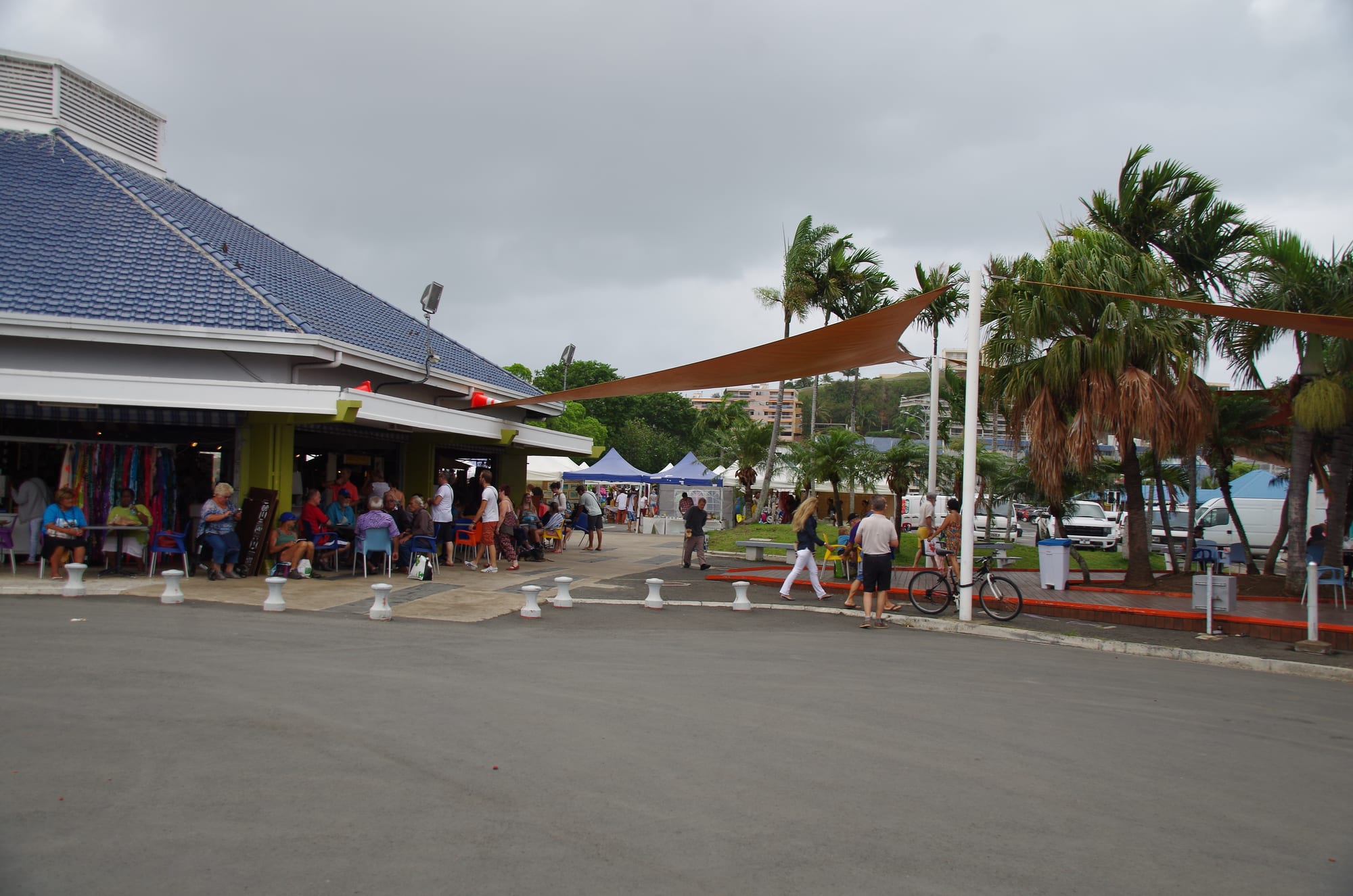 People sitting at tables and walking near a blue-roofed market pavilion beside palm trees in Noumea