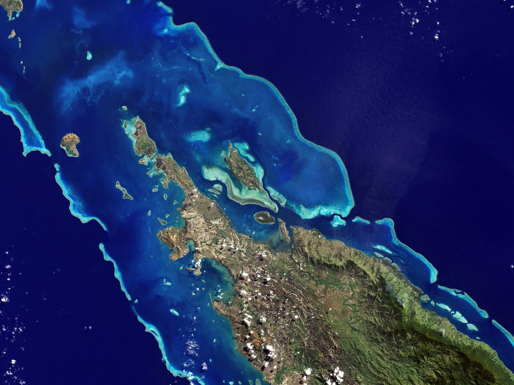 A satellite view of a large island ringed by turquoise lagoon and a long barrier reef