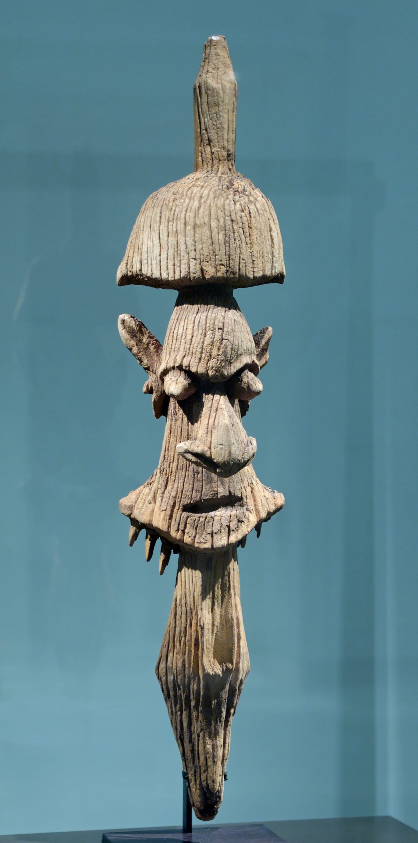 A weathered pale wooden carving of a stylised face with a large hooked nose, ear-like projections and a tall pointed crown, displayed against a blue-green wall