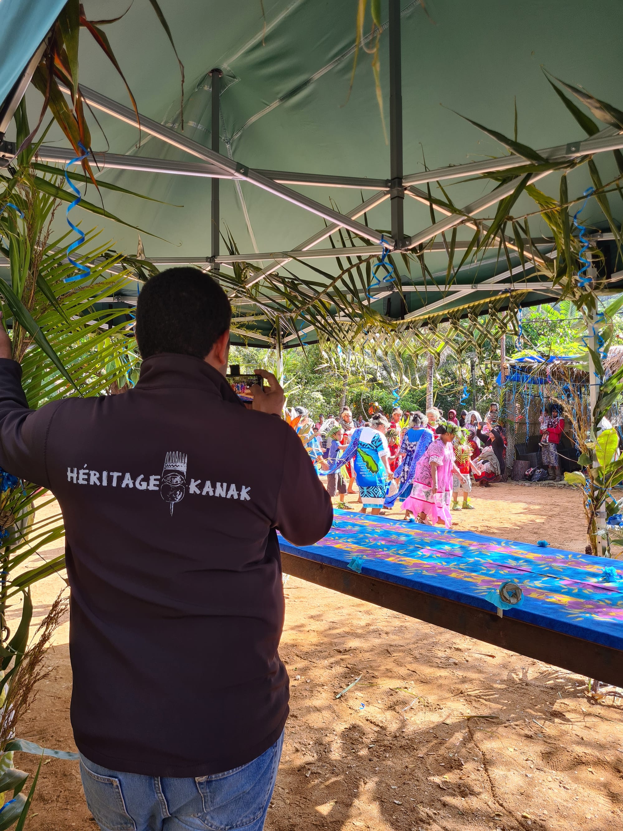 A man seen from behind in a dark jacket reading Héritage Kanak films a group of women in brightly coloured loose dresses dancing on bare earth beyond a palm-decorated shelter