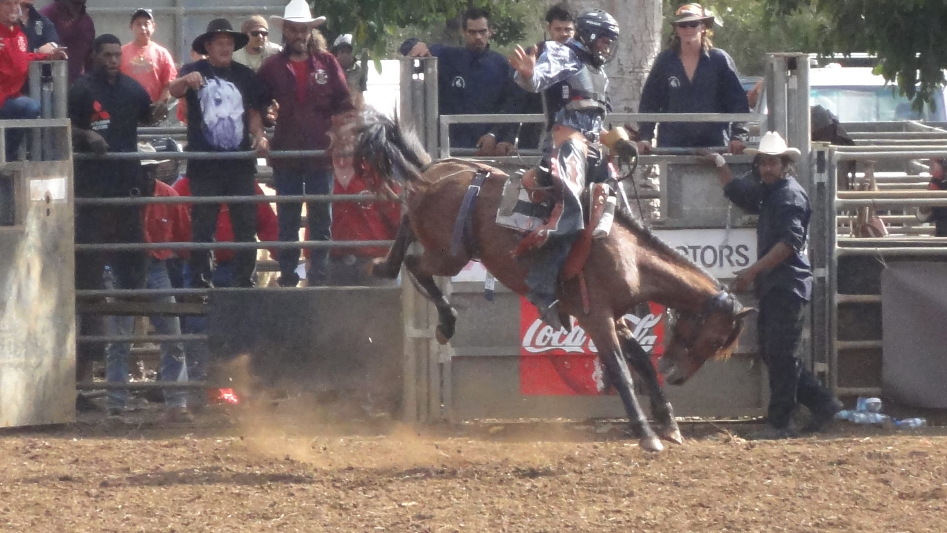 A rider in a helmet and protective vest clings to a bucking brown horse in a dusty arena, with spectators in wide-brimmed hats watching from behind metal rails
