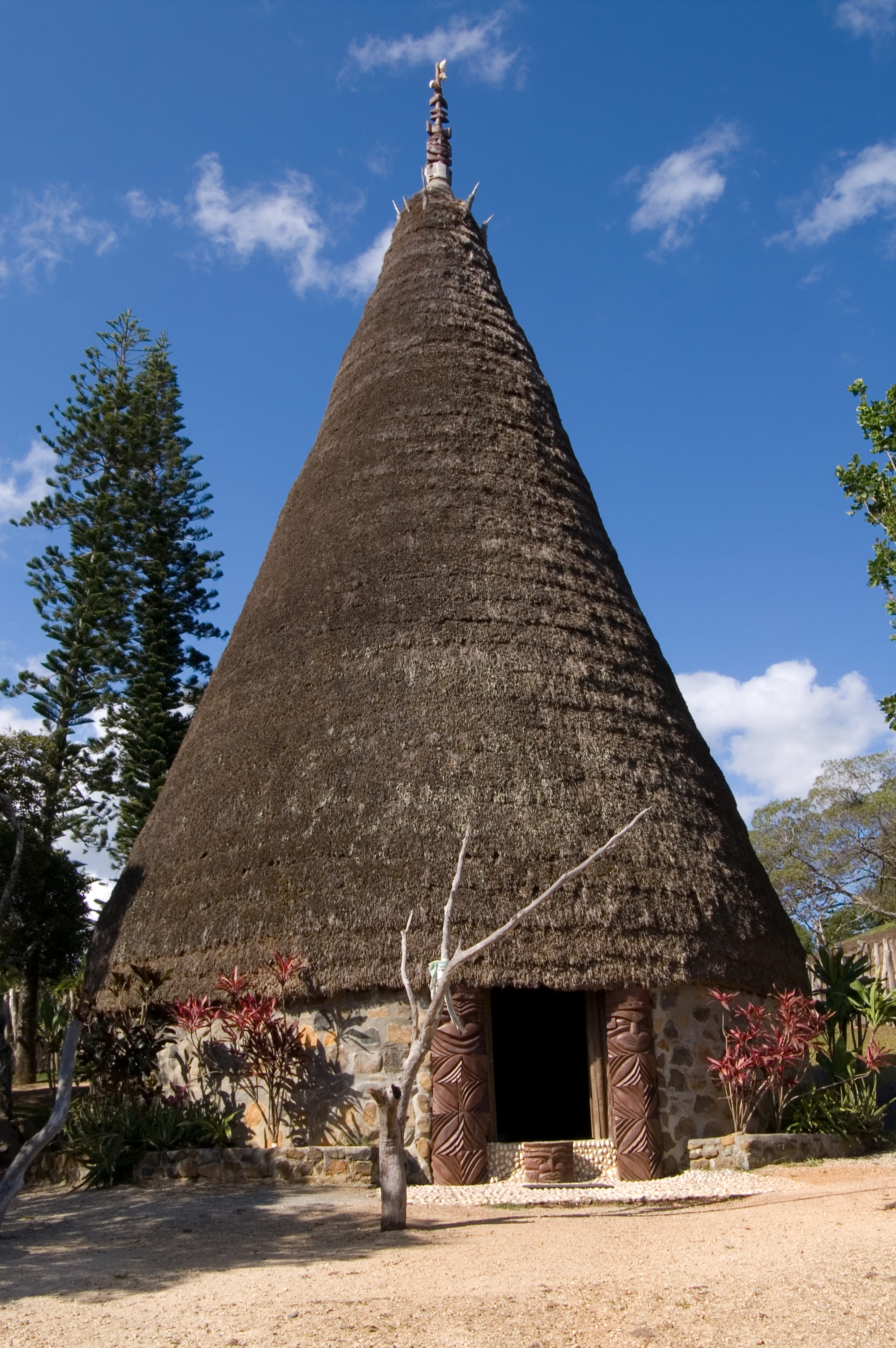 A tall conical Kanak great house with a thick thatched roof topped by a carved wooden spire, its doorway flanked by two carved wooden posts, under a blue sky
