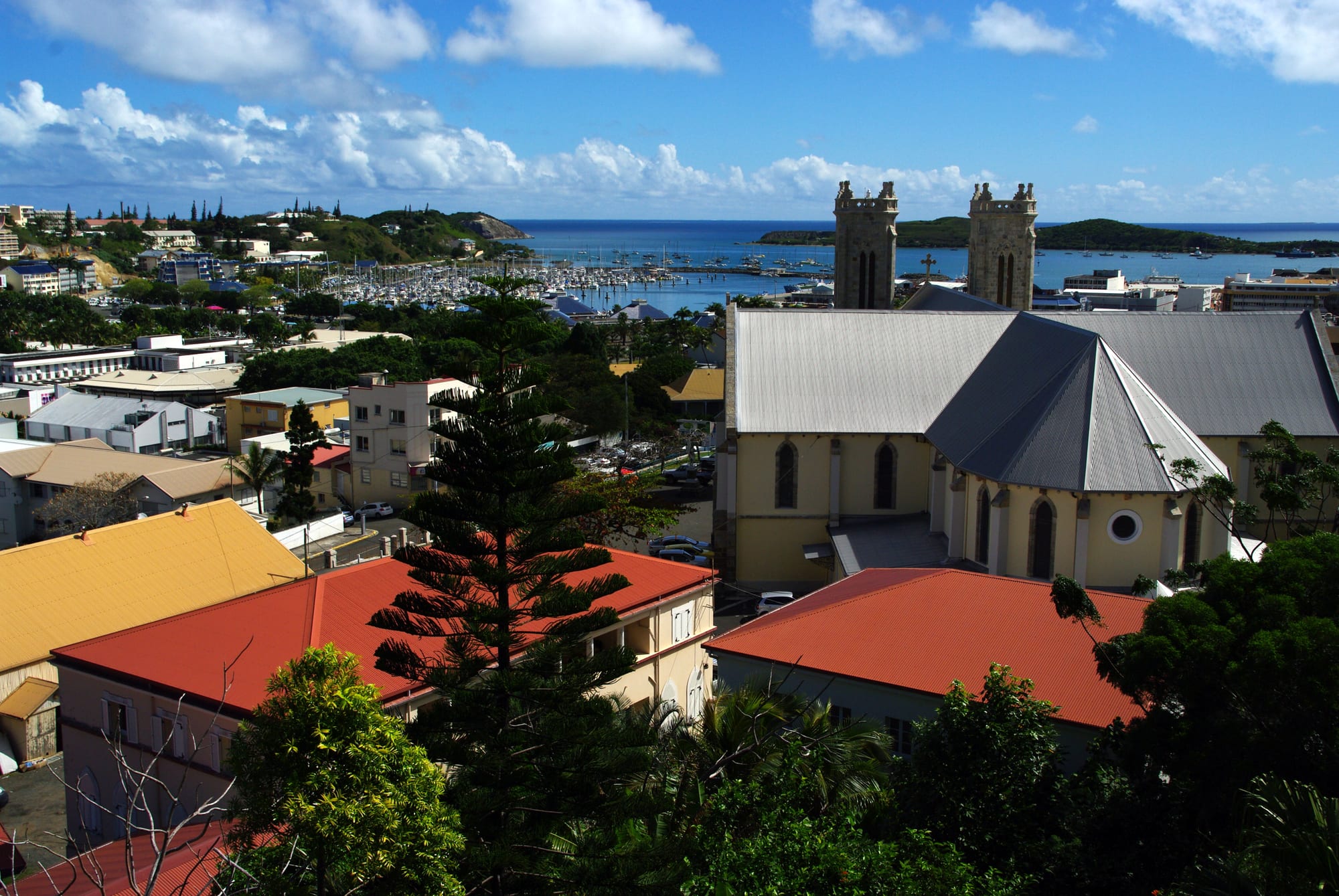 A view over the red-roofed city of Nouméa with a cathedral tower, a bay and forested hills beyond
