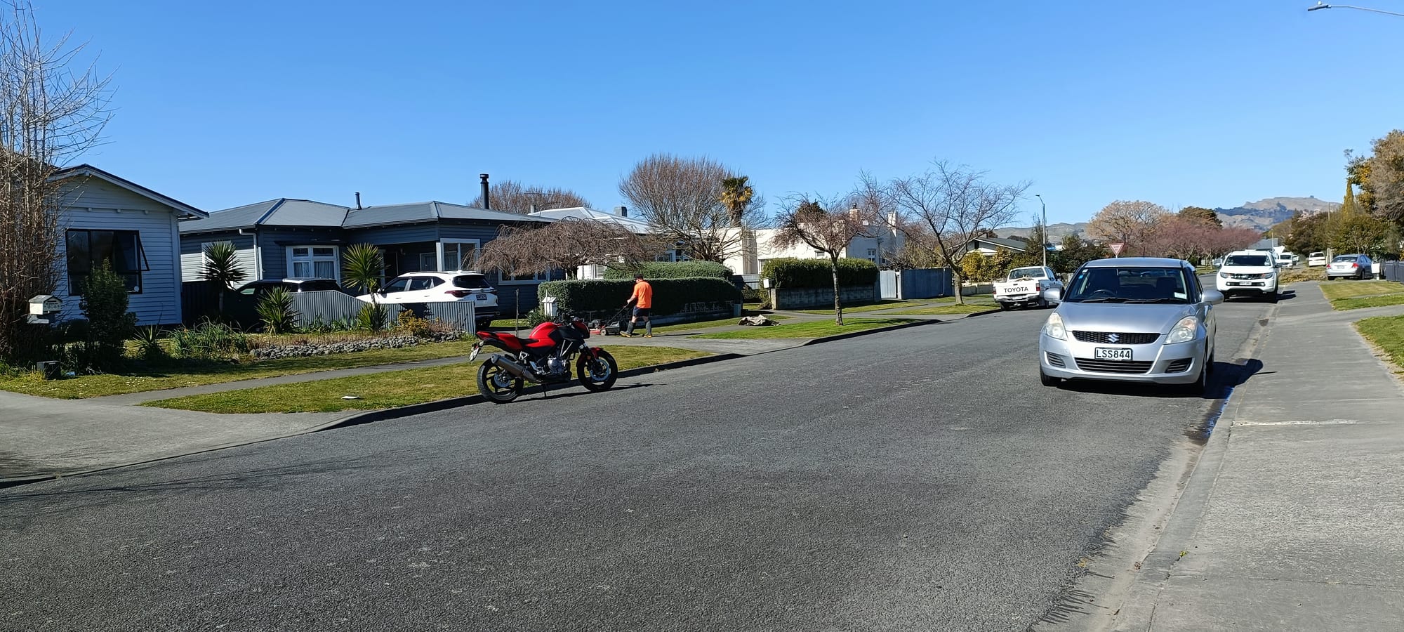 A suburban street of single-storey houses with front gardens, parked cars and a motorbike, a man mowing his lawn, and hills visible in the distance under a clear blue sky