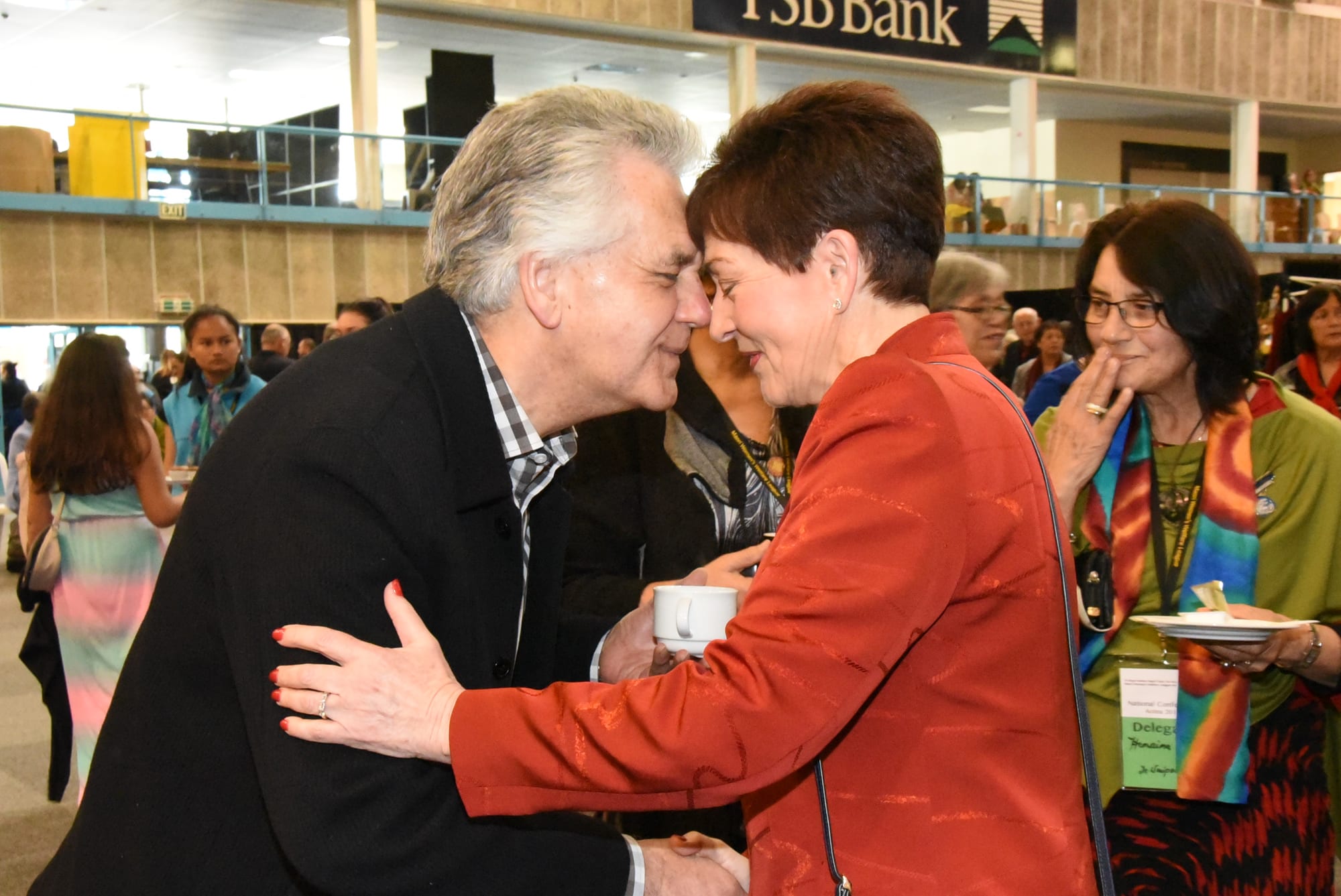 A grey-haired man and a woman in a red jacket lean towards each other with noses touching and hands clasped, as a woman in a colourful scarf looks on in a busy hall