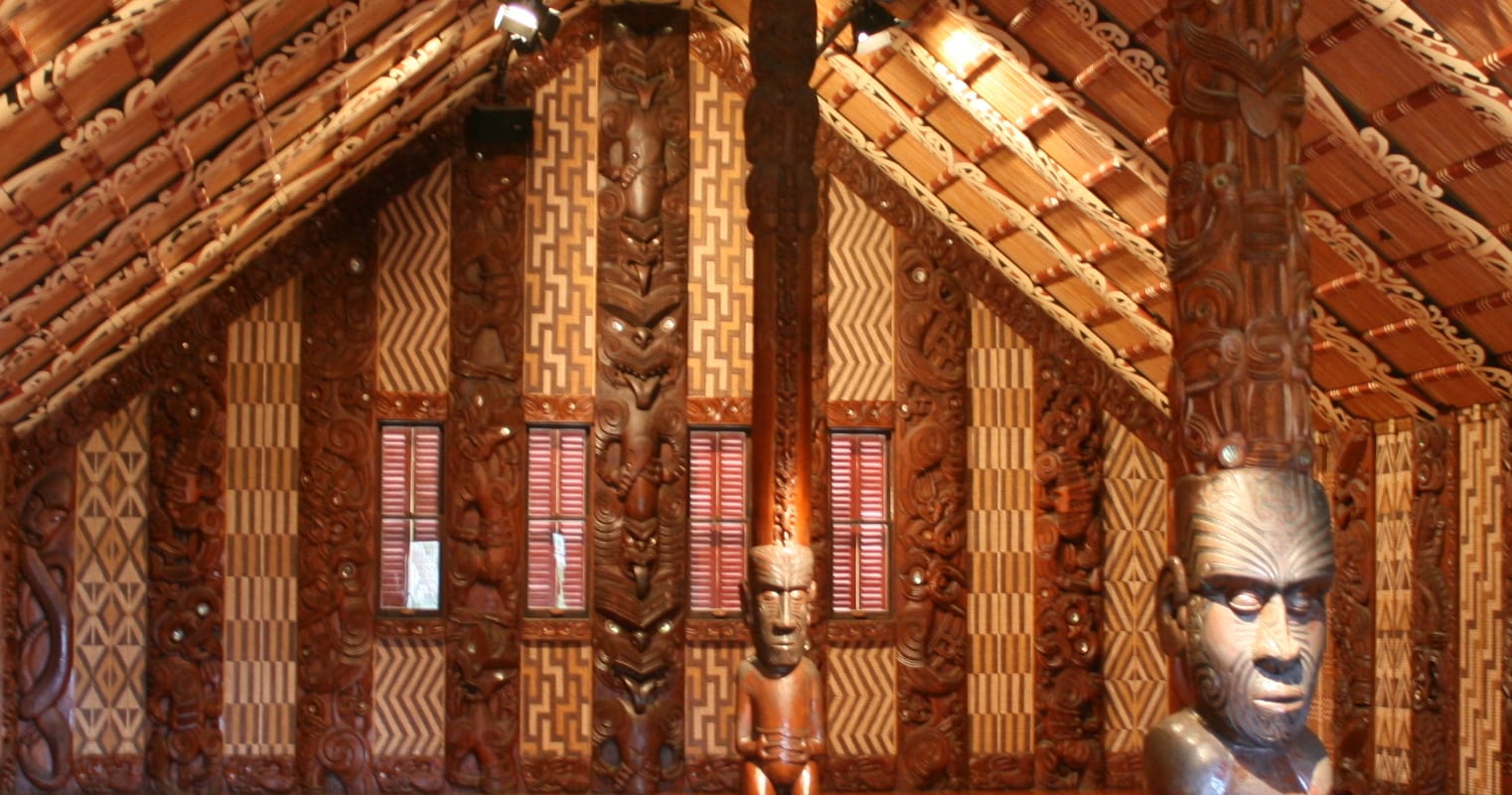 The interior of a meeting house with richly carved wooden wall panels and posts, woven lattice panels in geometric patterns, painted rafters and carved human figures in the foreground