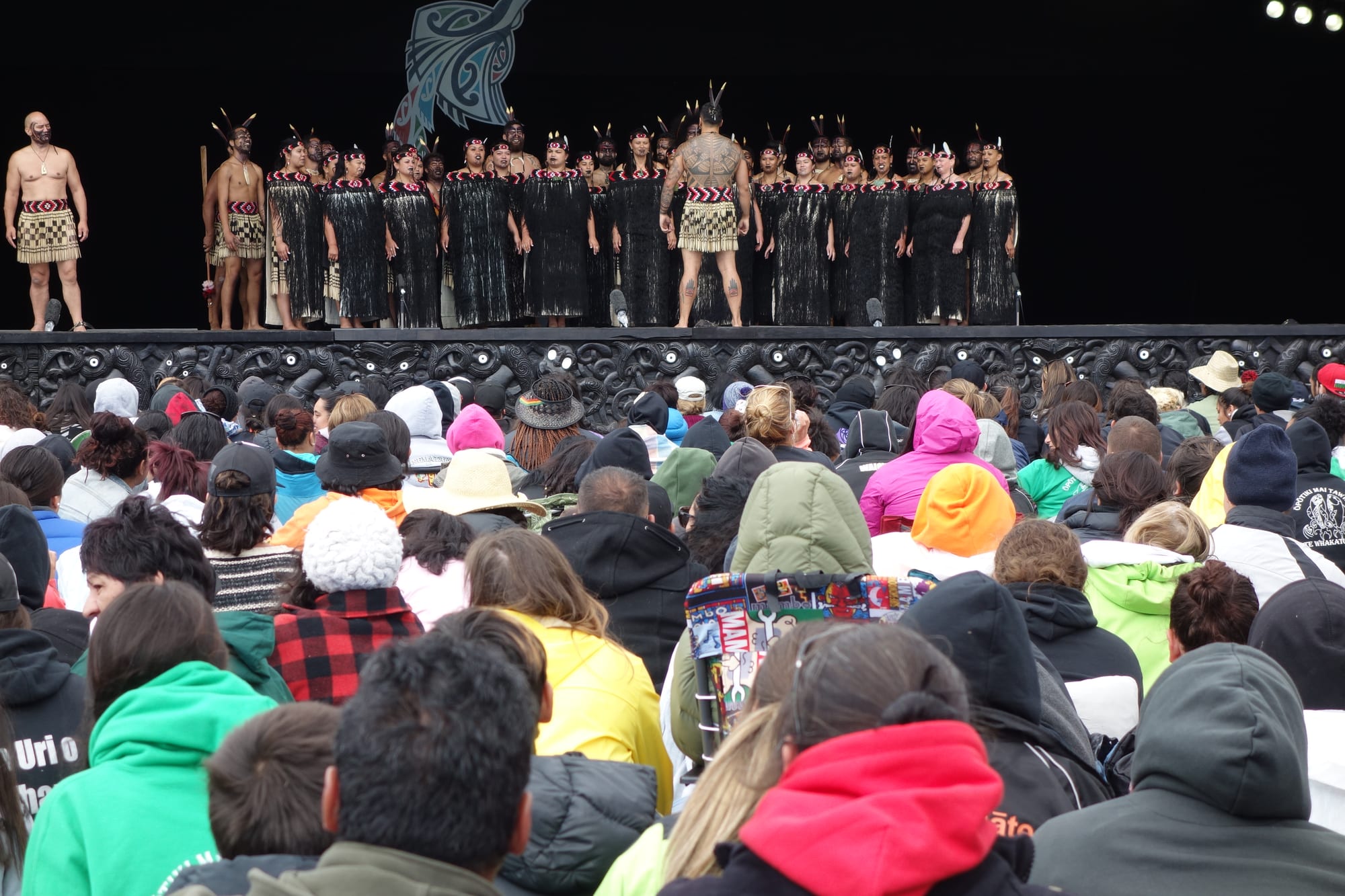 A large kapa haka group in black and red costumes with feathers in their hair stands on an outdoor stage with a carved black and white front, watched by a crowd in warm jackets and hats