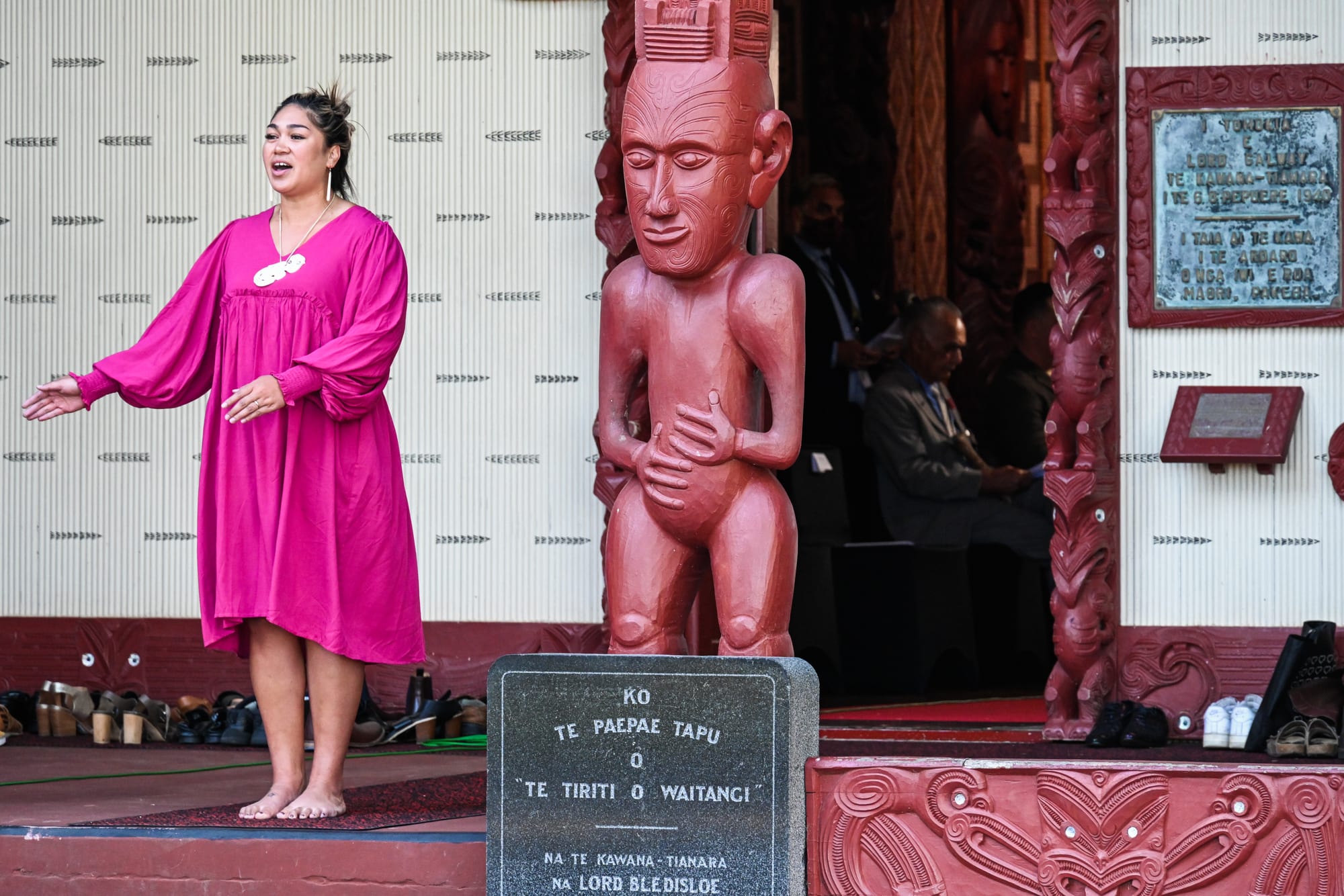 A woman in a pink dress stands barefoot on the porch of a carved meeting house, arm outstretched as she calls out, beside a tall red carved figure, with rows of shoes left along the edge of the porch