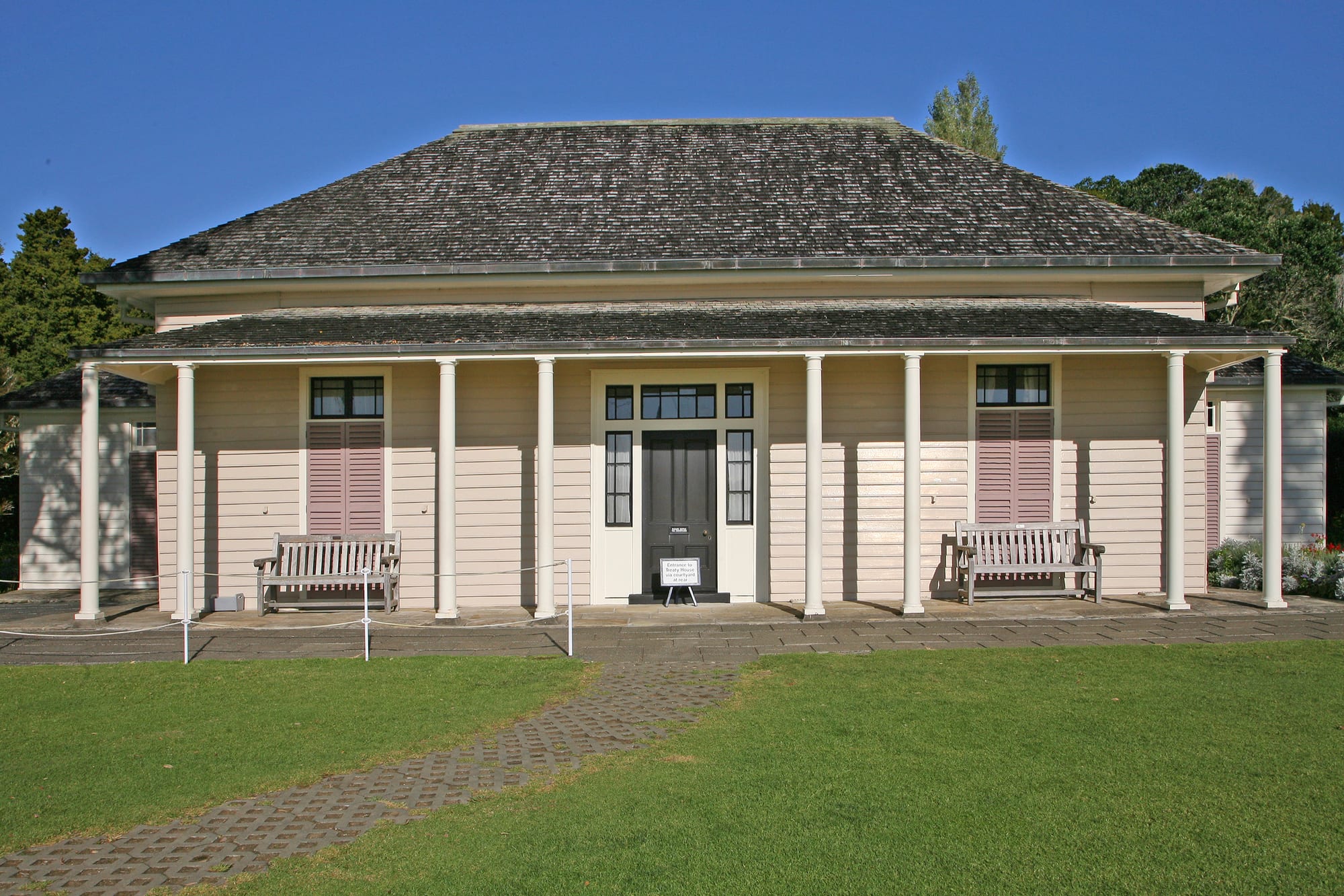 A white weatherboard colonial house with a wide verandah on a green lawn above a bay, with a flagstaff nearby