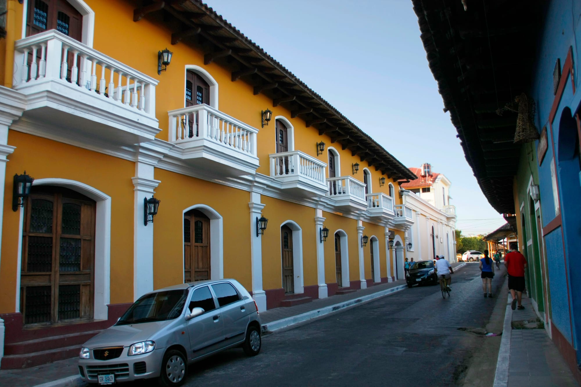 A row of single-storey colonial houses painted yellow with white balconies and arched doorways along a narrow street, cars parked outside