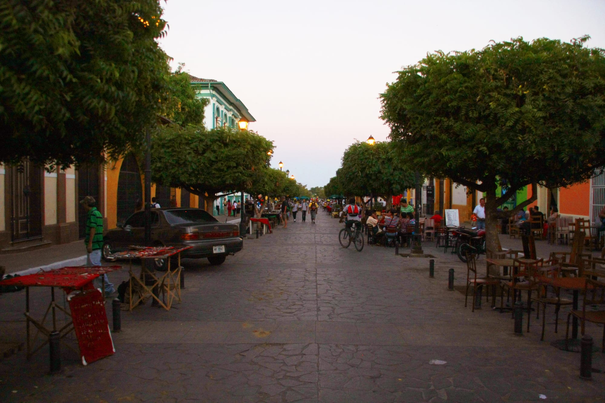 A tree-lined pedestrian street in Granada at dusk, lined with café tables, a cyclist riding past and people walking beneath string lights