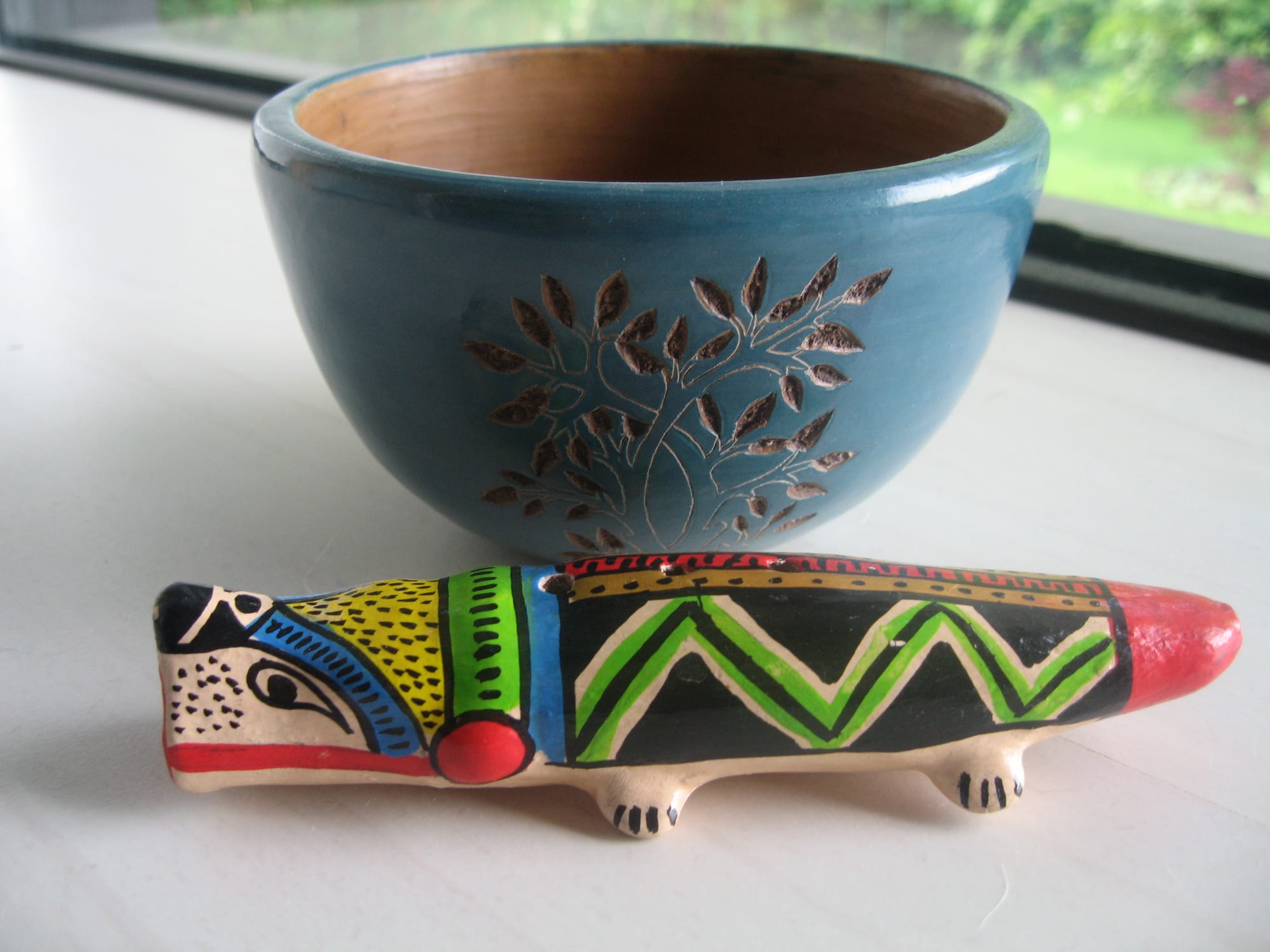 A blue-glazed ceramic bowl incised with a leafy tree design, behind a small clay flute shaped and painted like an animal with bright green, yellow and red patterns