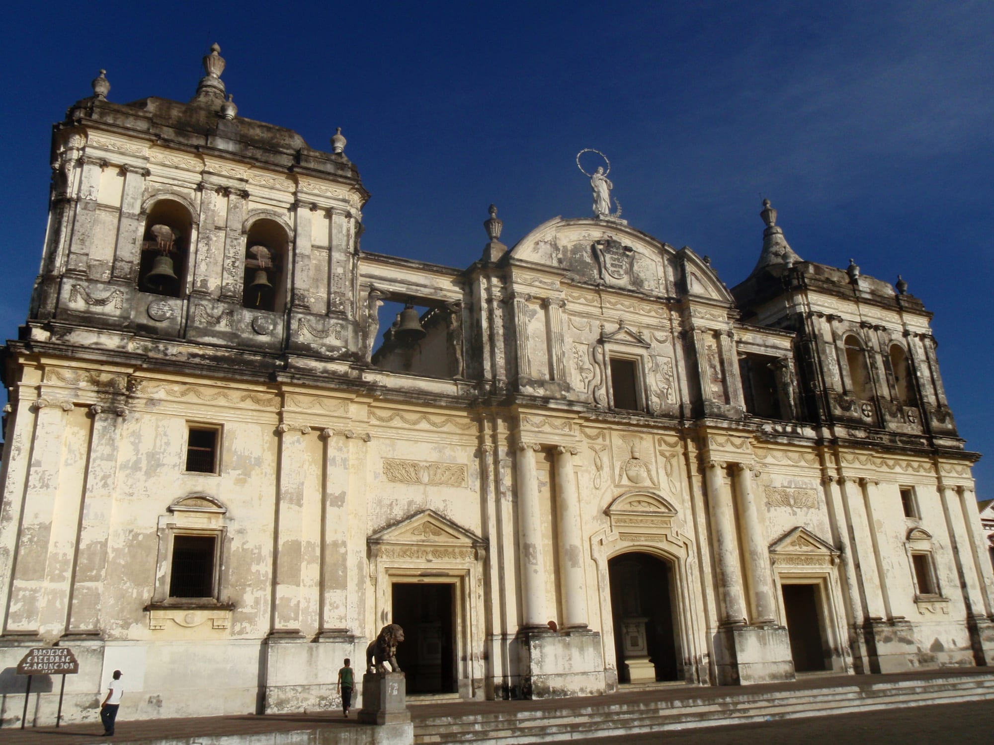 The broad, weathered white facade of a colonial cathedral with two bell towers, arched doorways and a statue on the central gable, under a deep blue sky