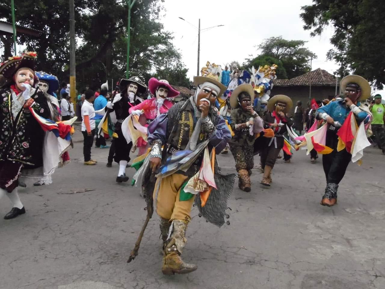 Dancers in pale painted wooden masks, wide hats and bright costumes with coloured scarves dance down a street, the leading figure carrying a staff