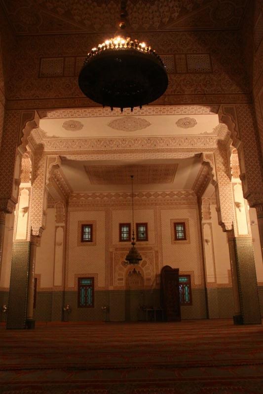 The tiled prayer hall of a mosque, with an ornate arched mihrab and a hanging chandelier