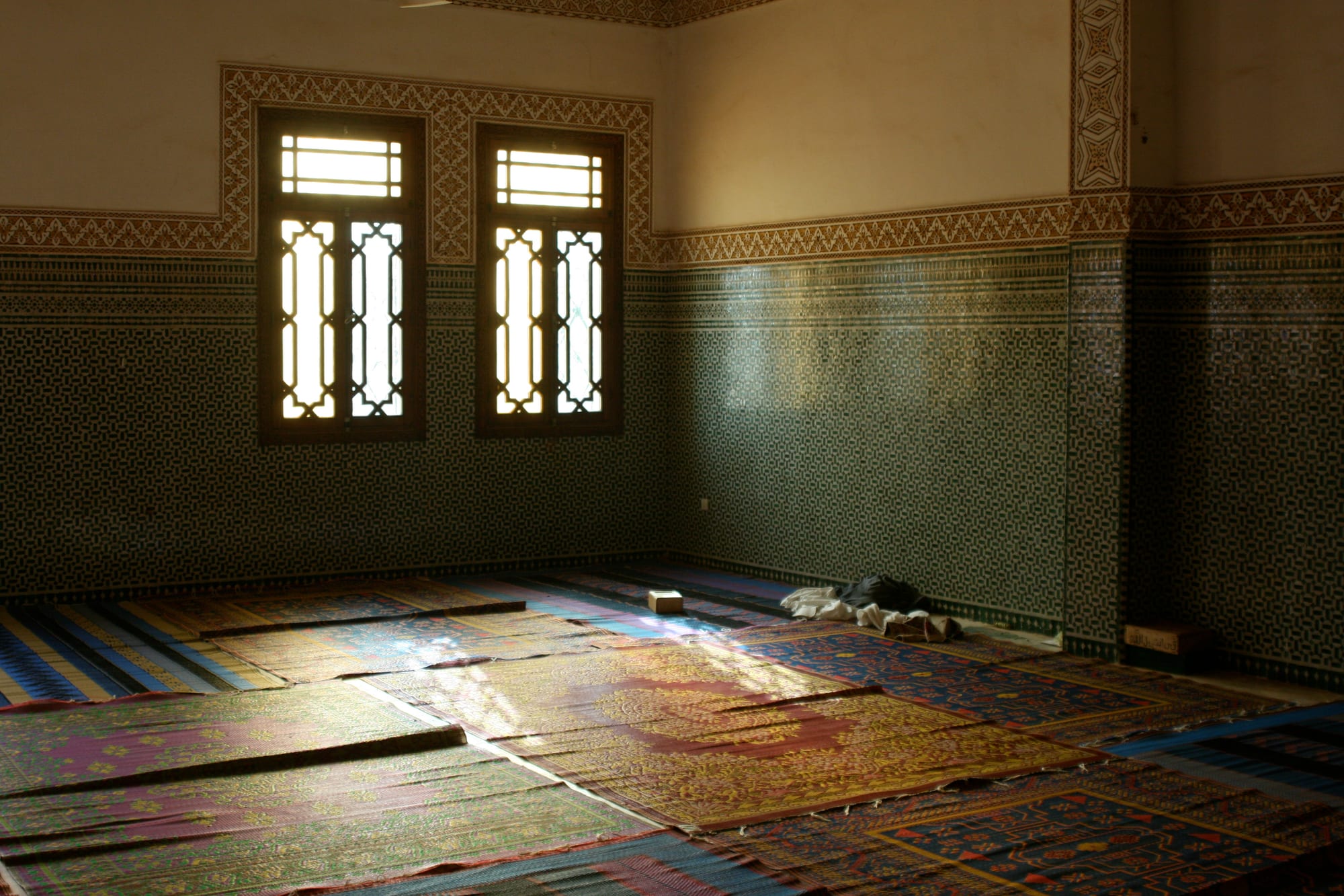 The interior of a mosque with patterned prayer mats covering the floor, walls of small green tiles and two latticed wooden windows letting in light