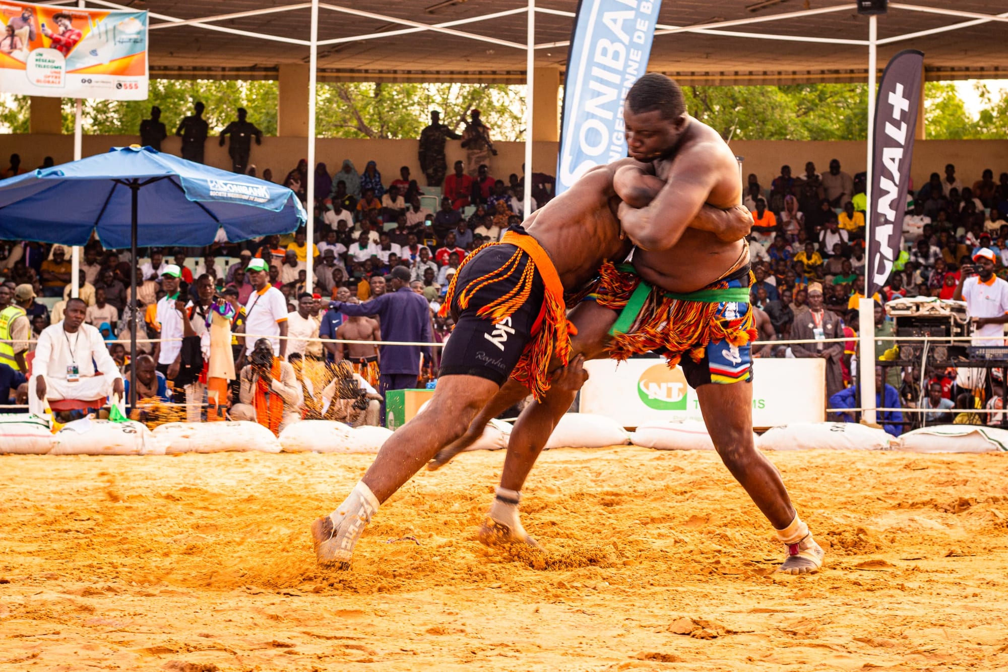 Two wrestlers in decorated loincloths and fringed belts grapple in a sand arena, one lifting the other, in front of a crowded grandstand