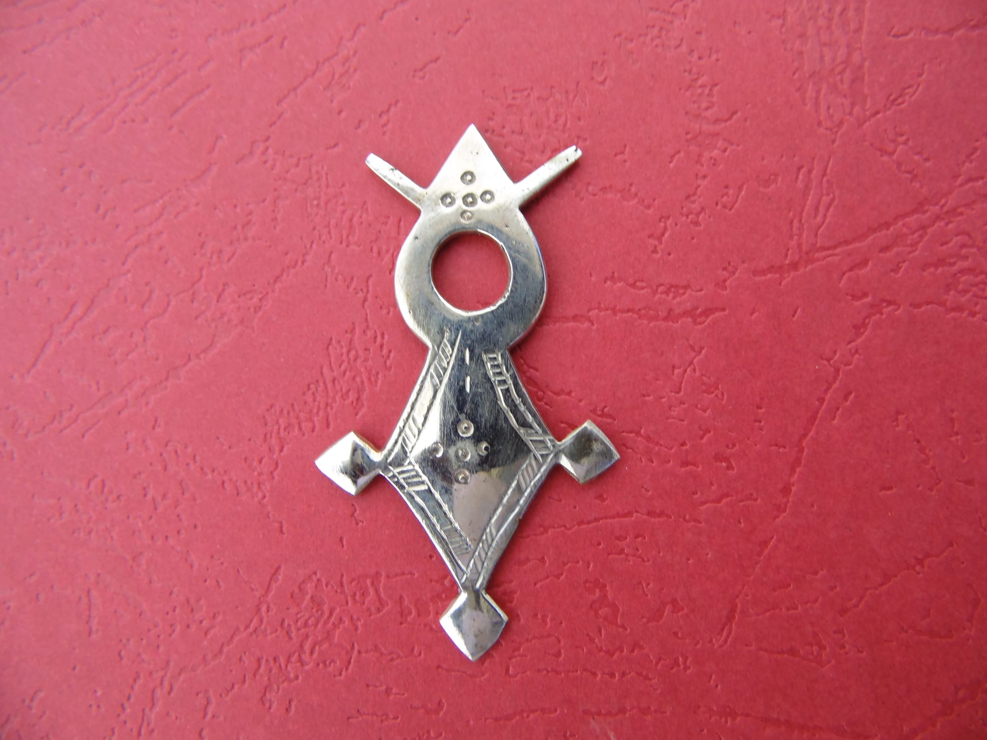 A polished silver pendant with a ring at its centre, a pointed top with two short arms, and a diamond-shaped lower body with engraved lines and dots, on a red background