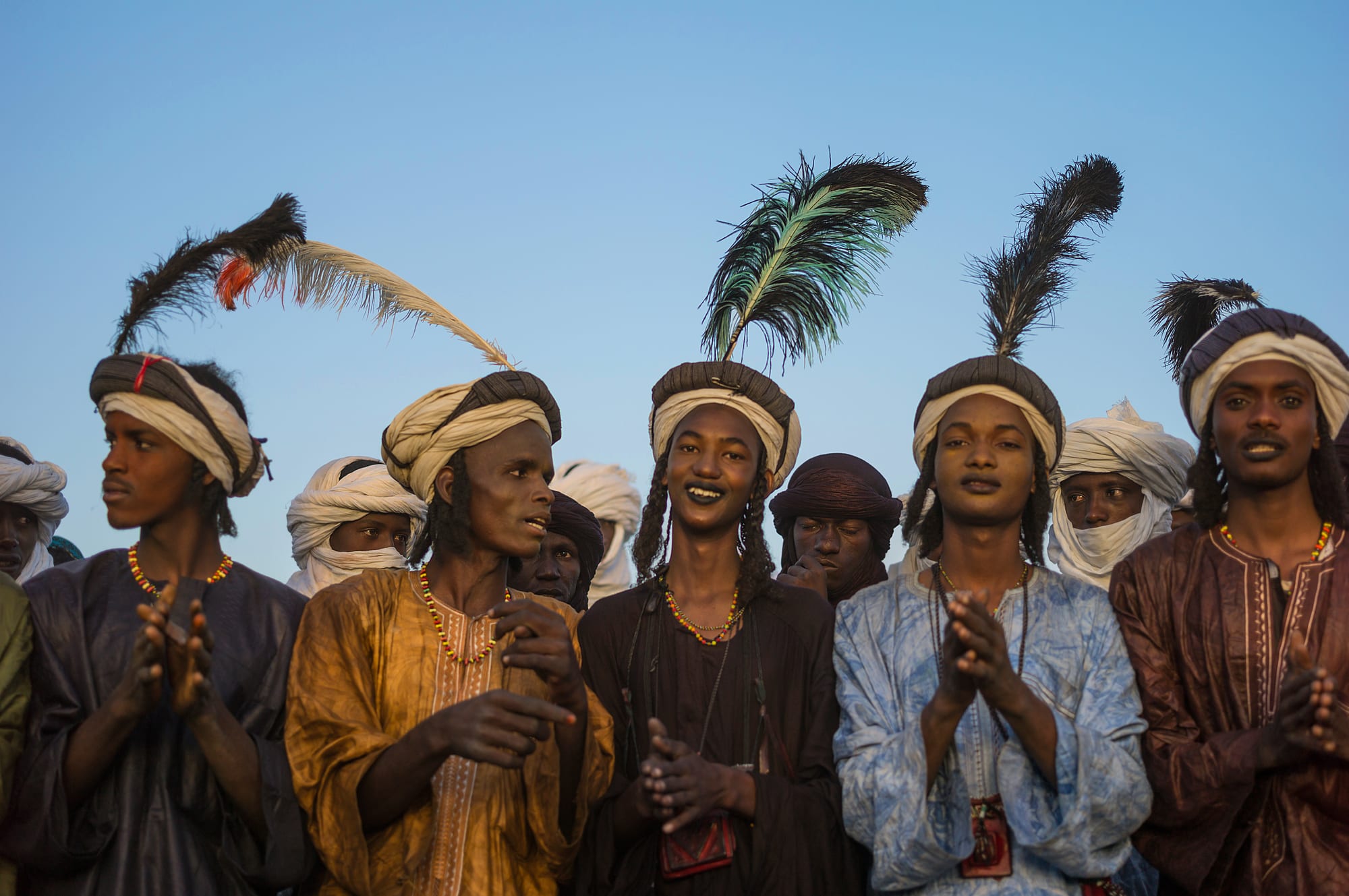 A line of young men in turbans topped with tall feathers, beaded necklaces and bright robes, some clapping and singing, against a clear blue sky
