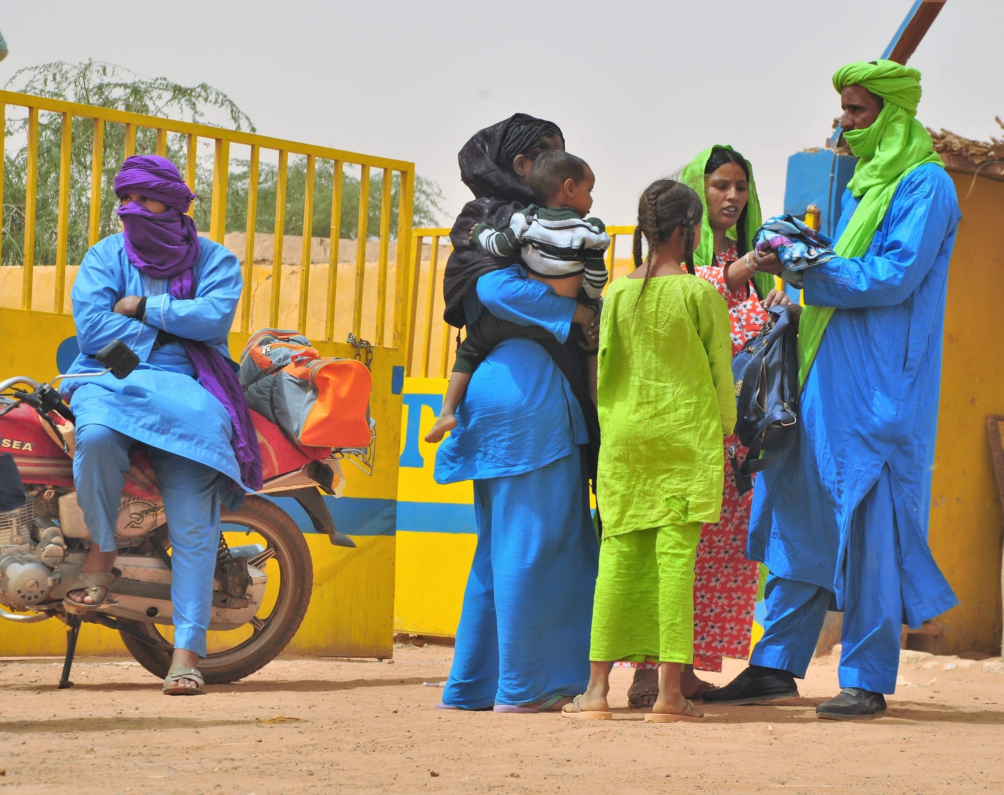 A family in brightly coloured robes, a woman carrying a small child on her back, gathered by a yellow gate next to a parked motorbike