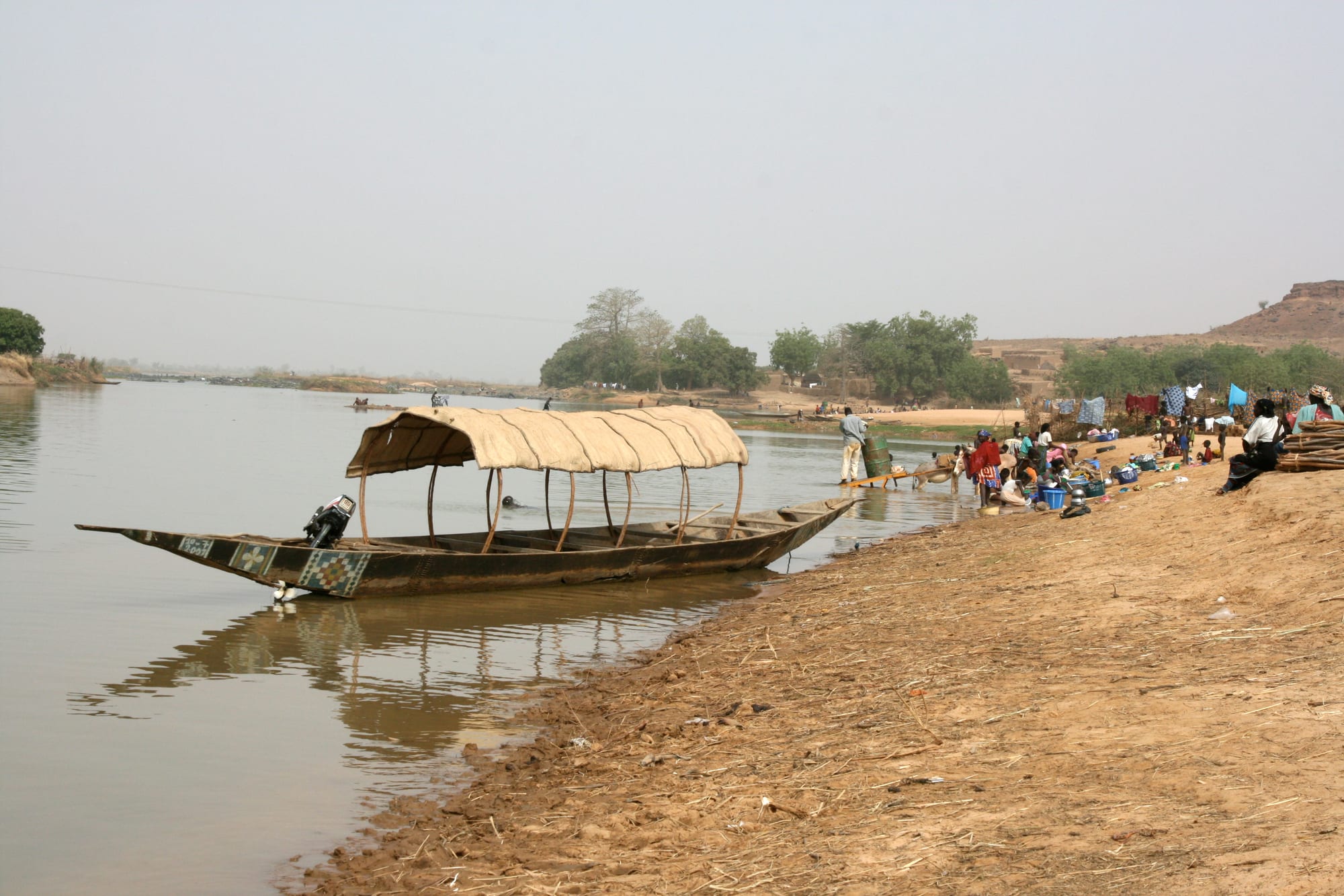 A wooden river boat with a woven canopy moored on a muddy riverbank, with people washing clothes and gathering water further along the shore
