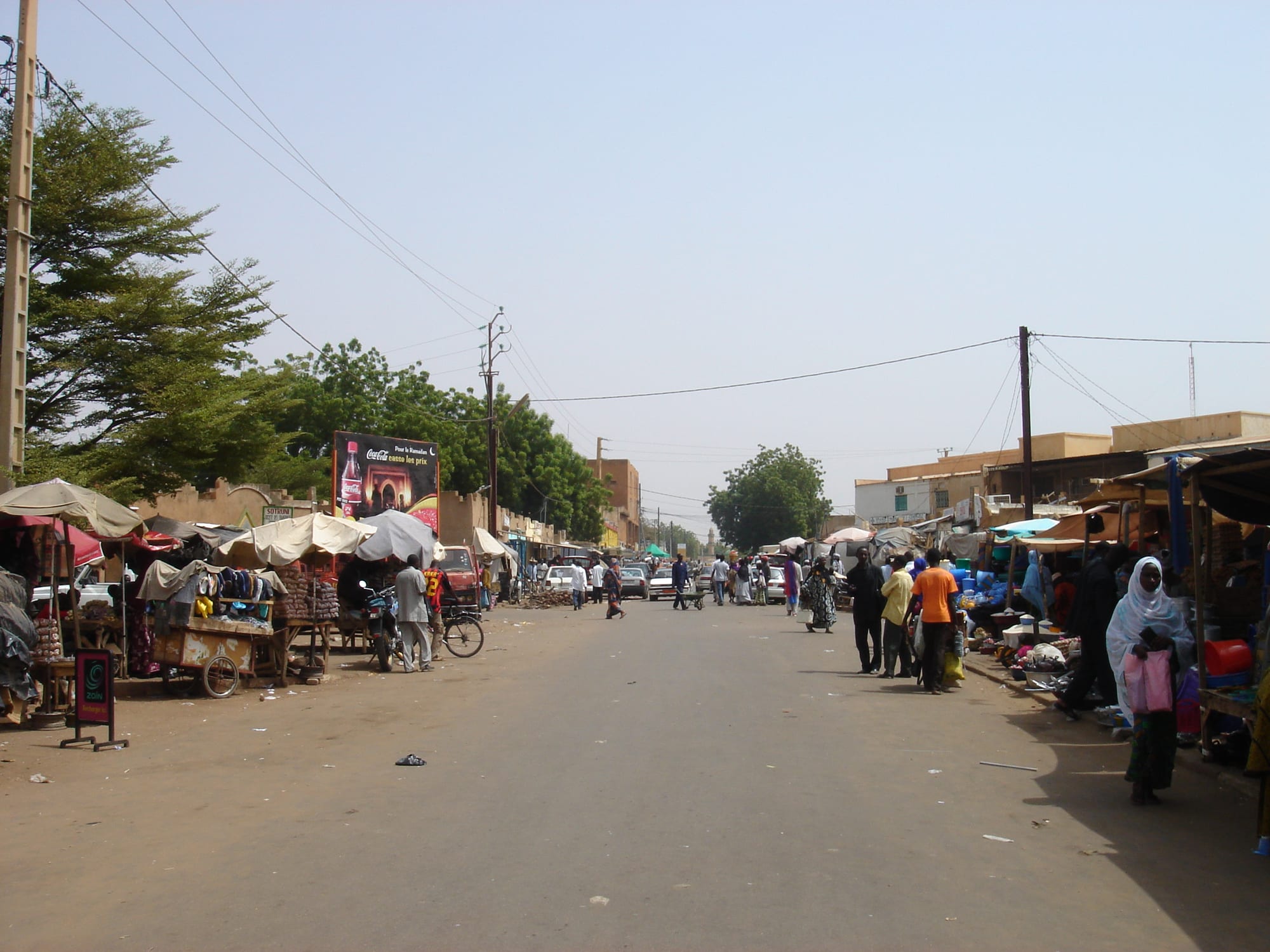 A wide street in Niamey lined with market stalls and umbrellas on both sides, with people walking and a mosque minaret visible in the distance