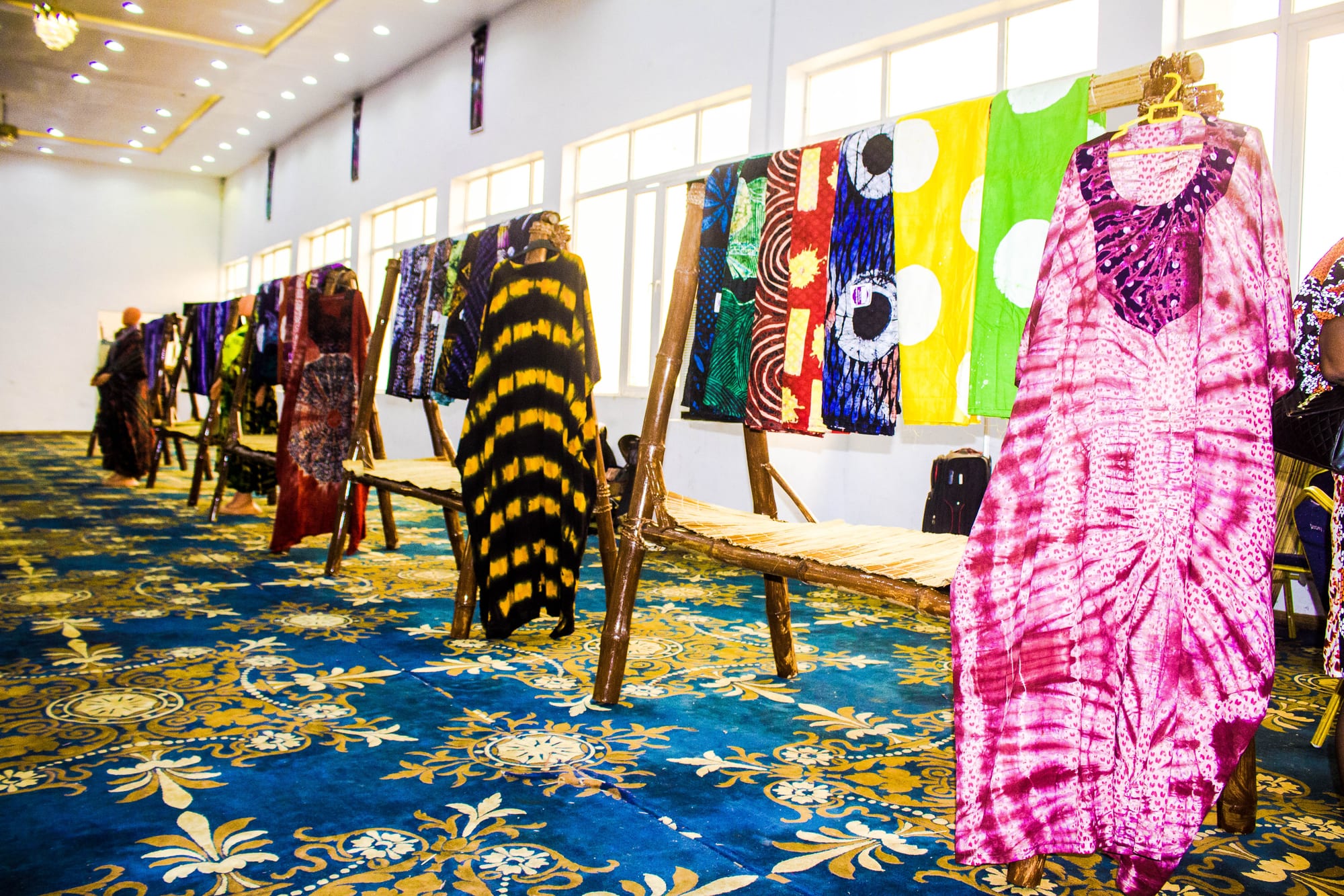 Tie-dyed and resist-patterned cloths in pink, yellow, blue, green and black hang from wooden stands in a bright hall with a patterned carpet