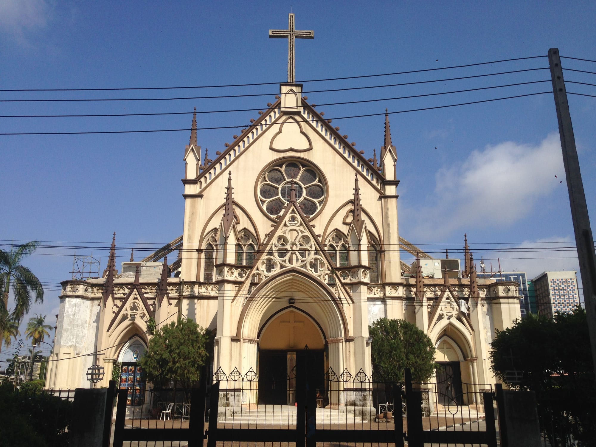 The cream-coloured facade of a church with pointed arches, spires, a large rose window and a tall cross on top, behind a black iron gate under a blue sky