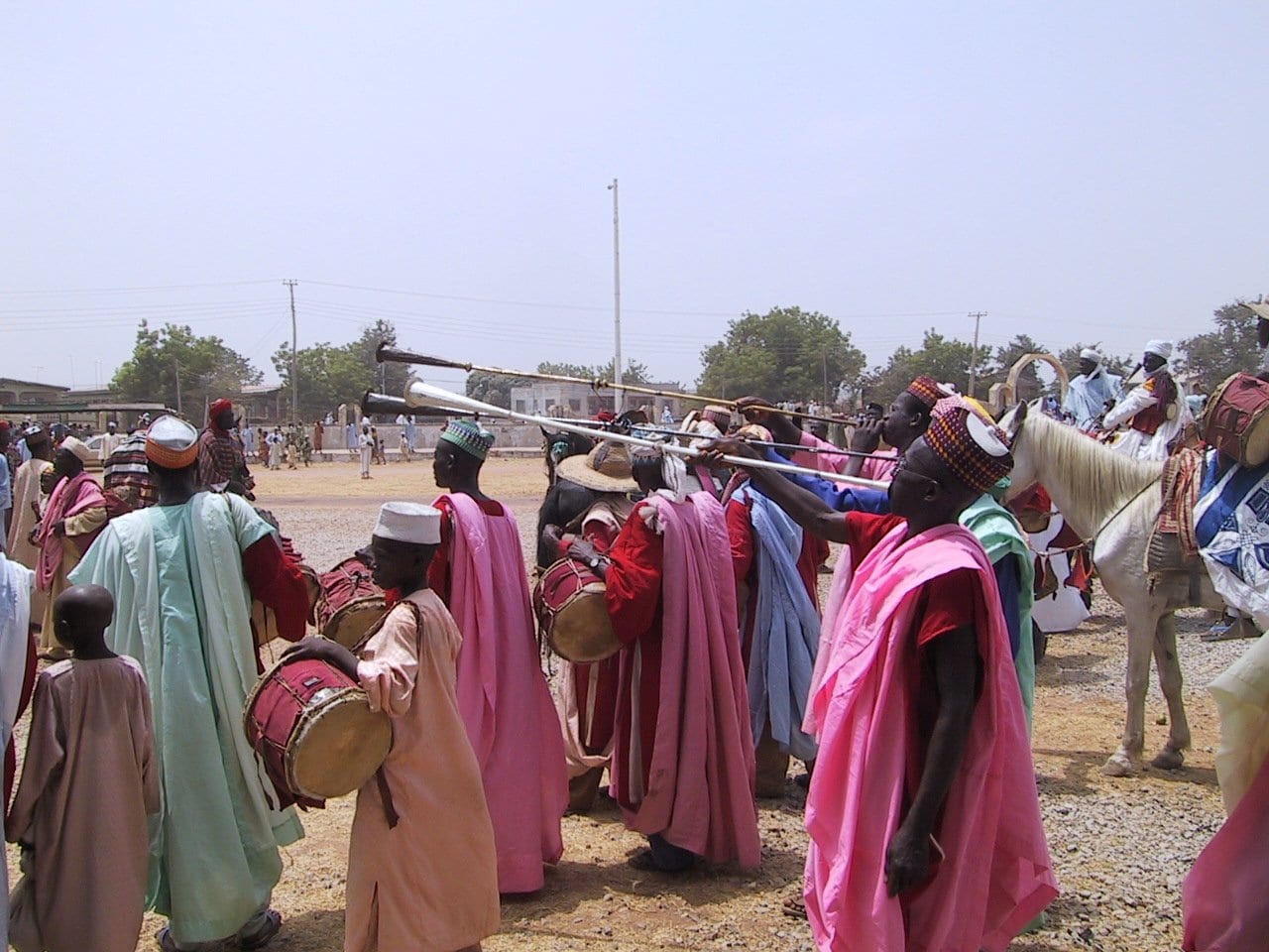 Men in long pink, red and pale green robes and embroidered caps blow long metal horns and carry drums beside a white horse on open, dusty ground