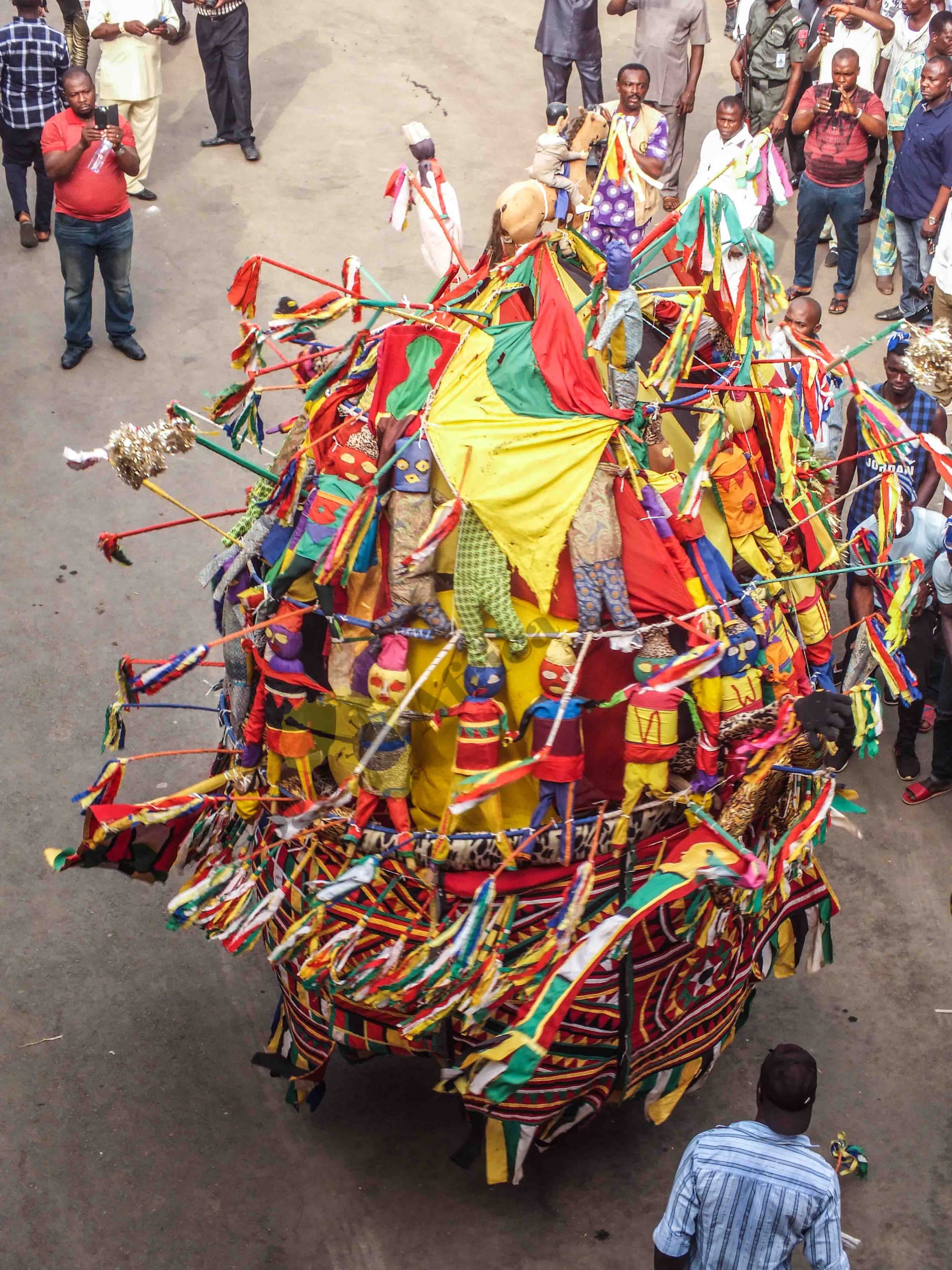 Seen from above, a towering masquerade costume of brightly coloured cloth panels, streamers and small carved figures moves along a road, watched by a crowd of men, some filming on their phones